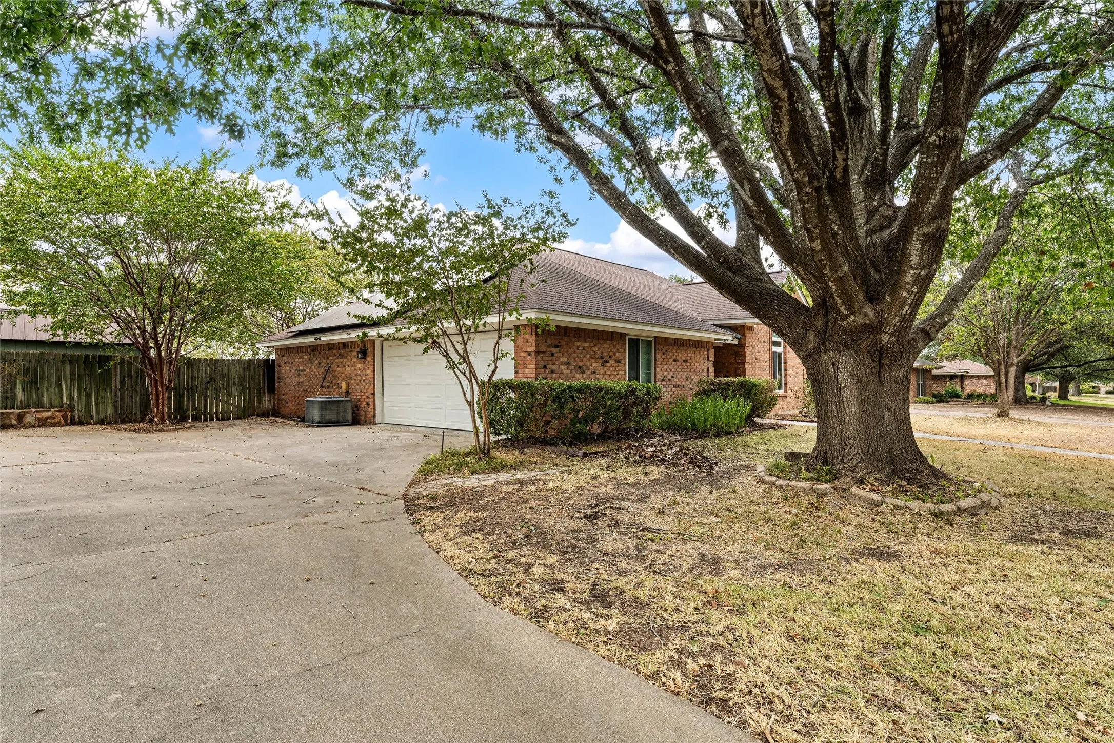 Brick exterior with an attached garage, paved driveway, mature trees, and established landscaping