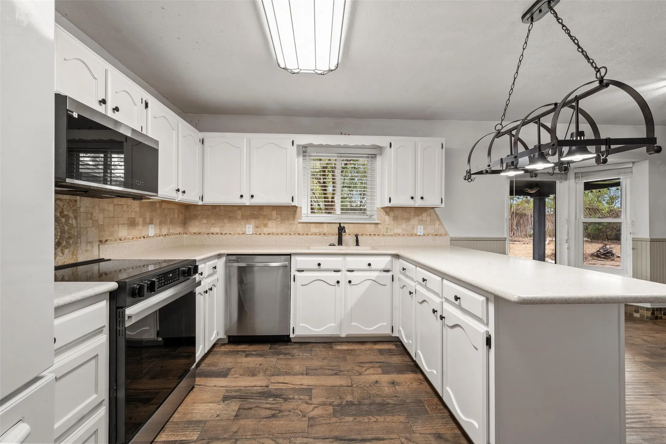 White cabinetry with black hardware, stainless steel appliances, wood-finish flooring, light-toned countertops, and a modern linear chandelier