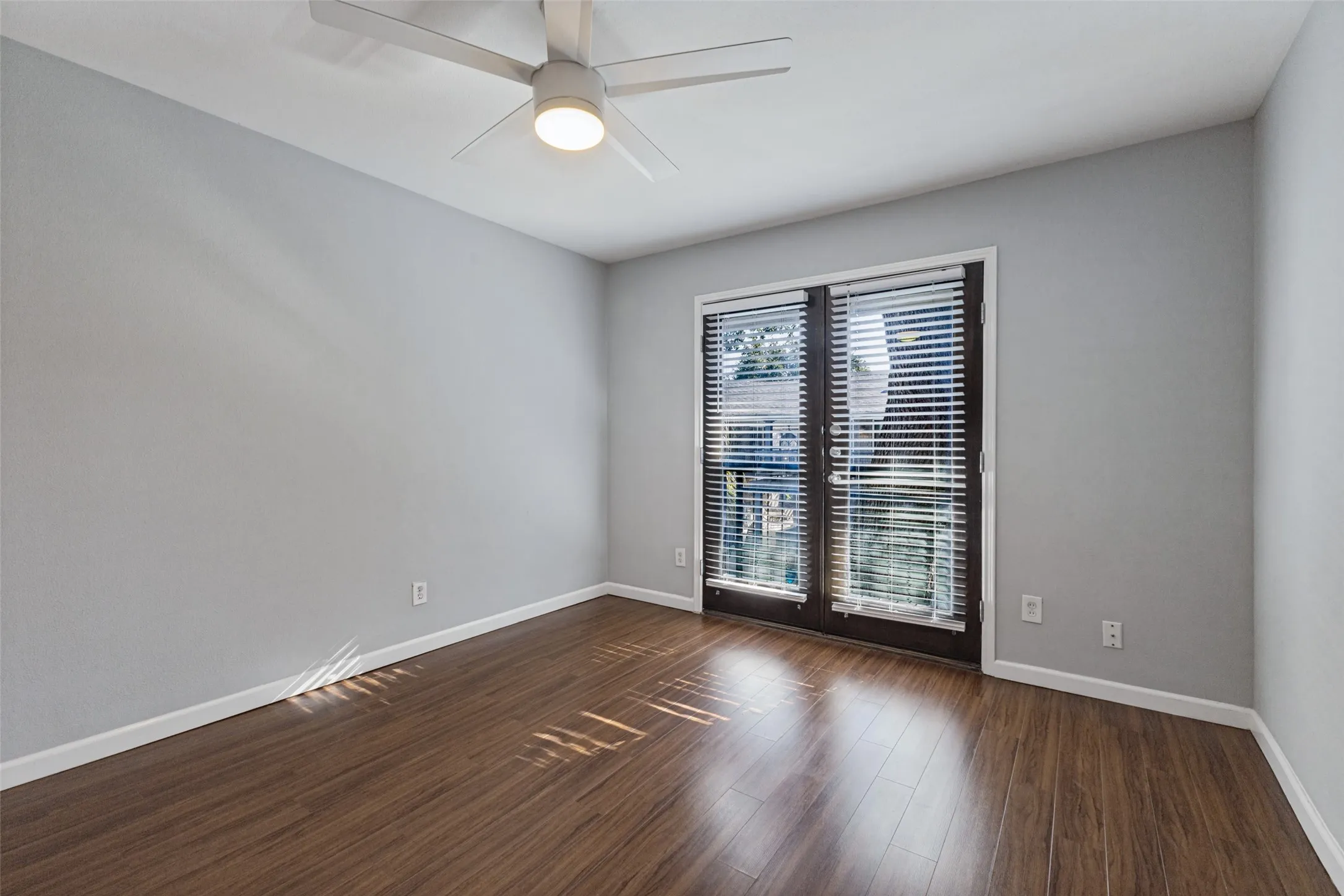 Wood-finish flooring, light gray wall finish, white ceiling fan with integrated lighting, and dark-framed double doors with horizontal blinds