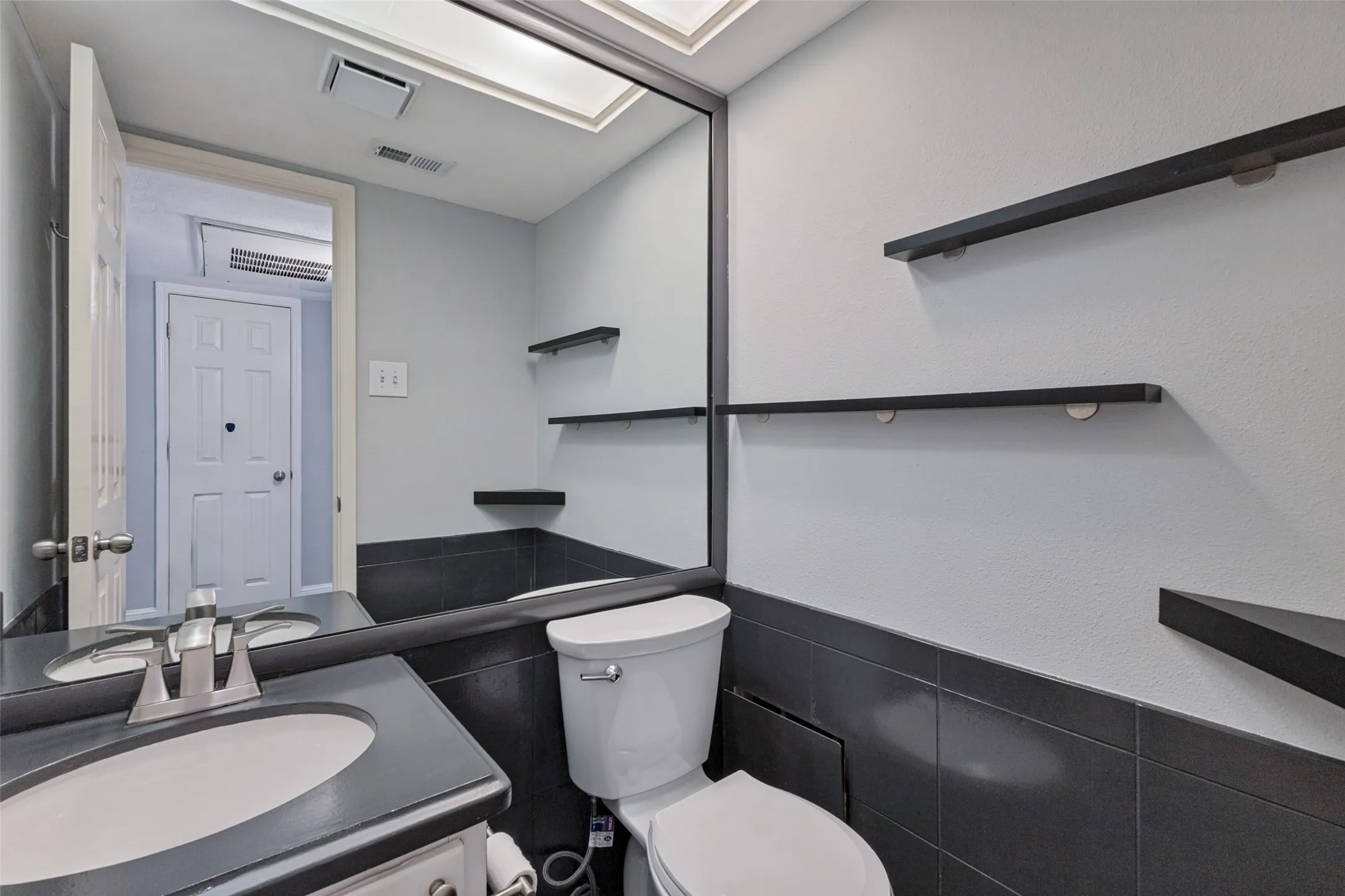Recessed lighting, wood-finish floating shelves, full-width vanity mirror, compact toilet, and a vanity with an integrated sink