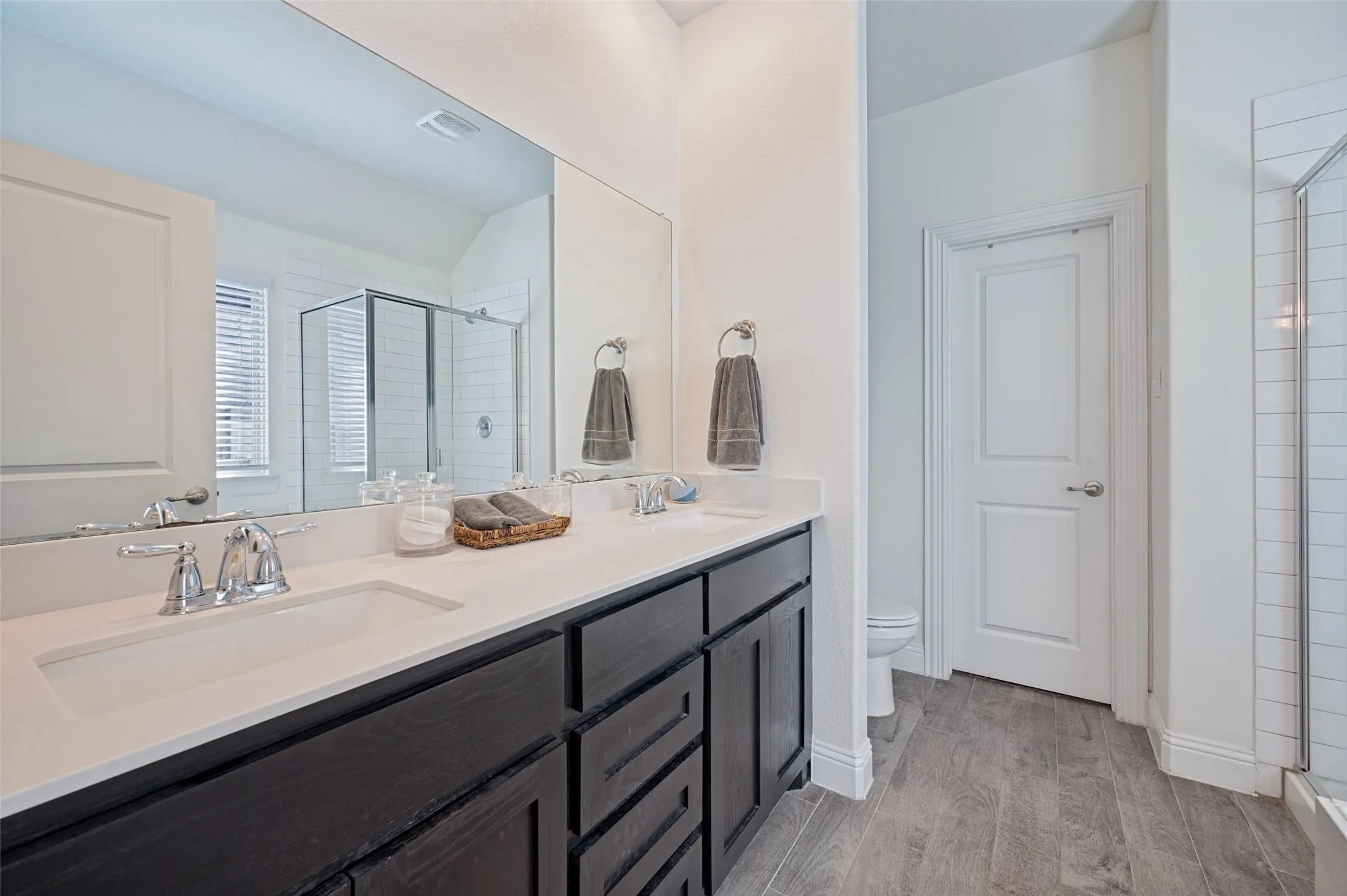 Dual vanity with chrome fixtures and integrated sinks, extensive mirror, dark wood-finish cabinetry, white countertop, and wood-finish tile flooring