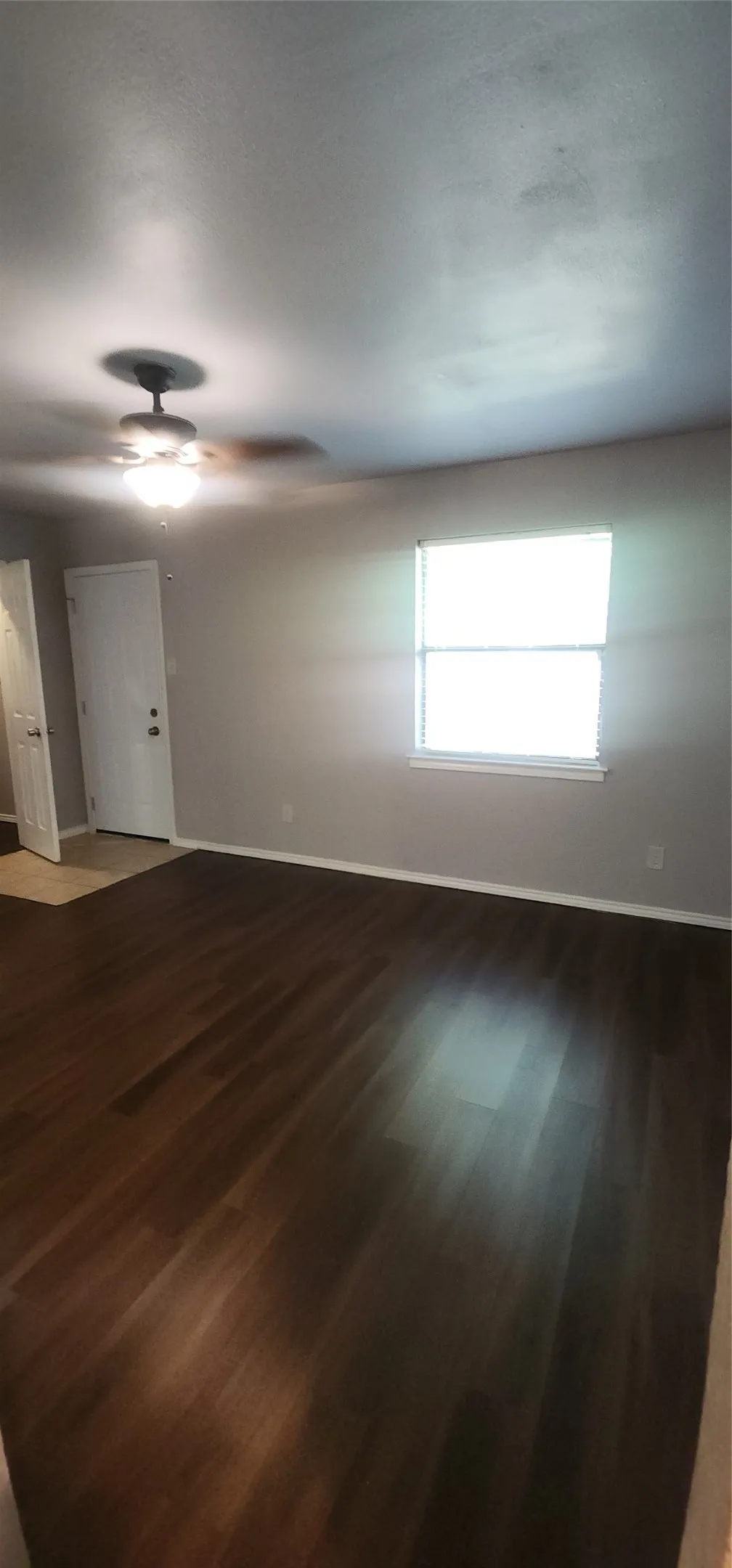 Dark wood-finish flooring, neutral wall paint, ceiling fan with integrated lighting, and a window with blinds