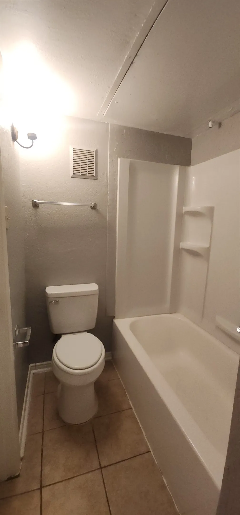Bathroom featuring a white toilet, white bathtub with a shower surround, tiled flooring, and a wall-mounted light fixture
