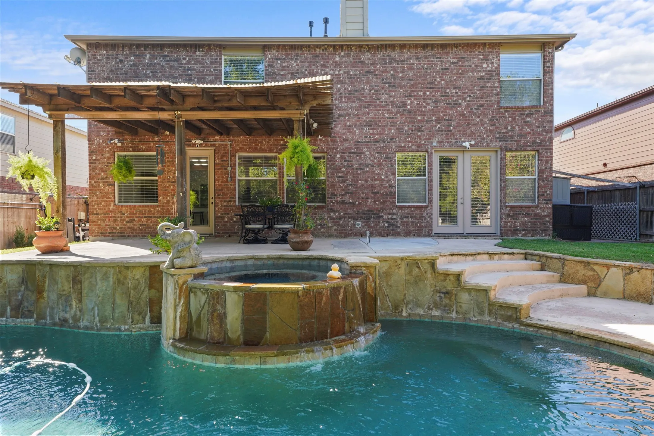 Brick exterior with a covered patio, integrated spa with waterfall feature, stone-clad pool surround, and curved stone steps