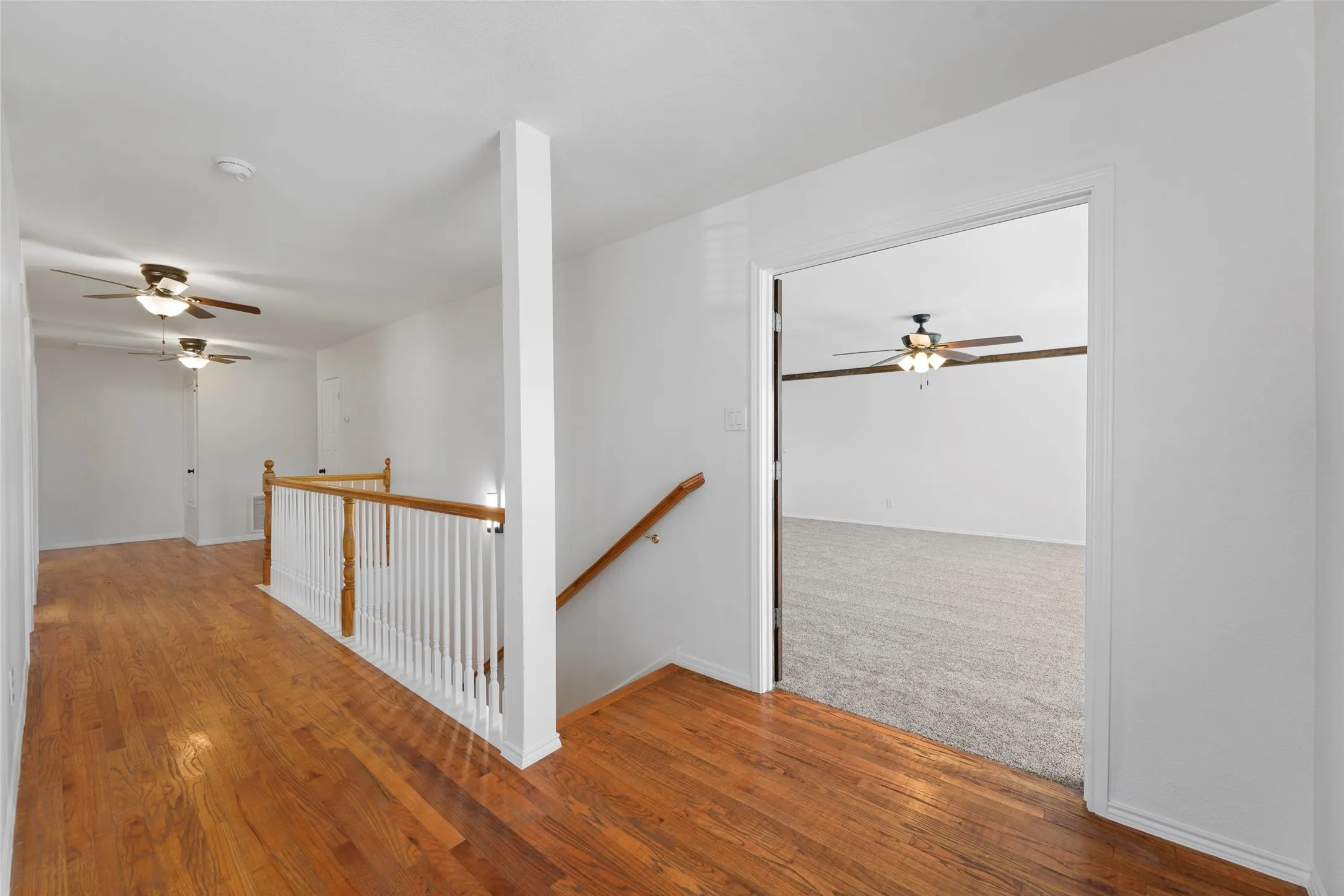 Hardwood flooring, white painted walls, white railing with wood-finish top rail, ceiling fans, and carpeted​​‌​​​​‌​​‌‌​‌‌​​​‌‌​‌​‌​‌​​​‌​​ room
