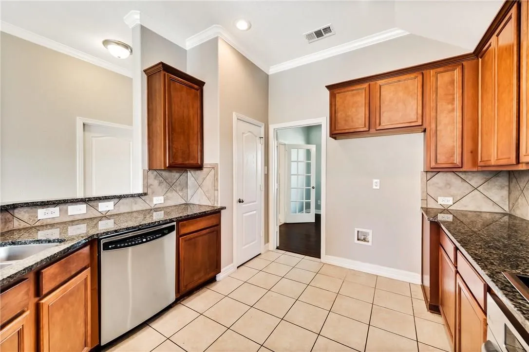 Kitchen featuring tile flooring, granite countertops, wood cabinetry, a stainless steel dishwasher, and a tile backsplash
