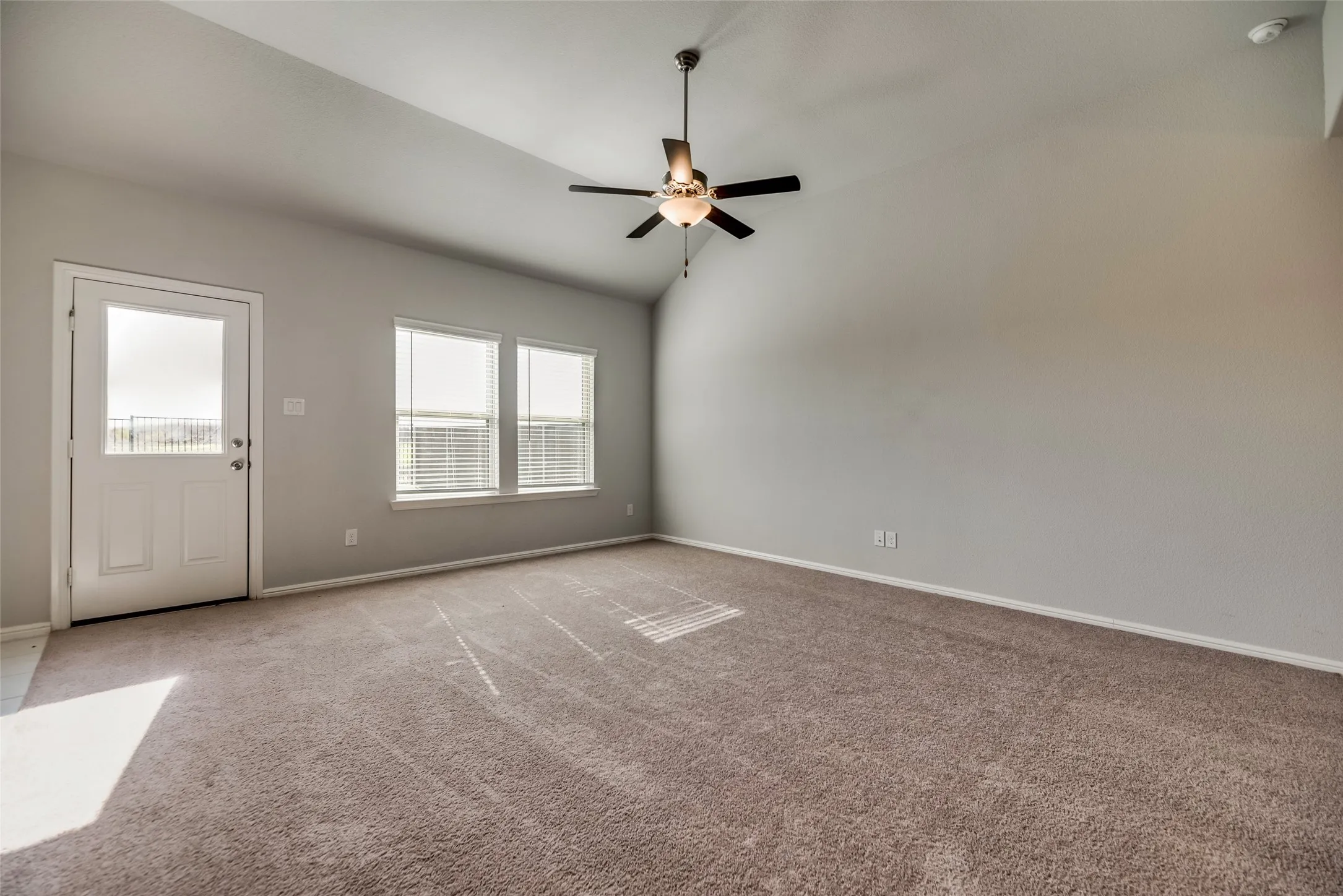 Spacious room featuring vaulted ceilings, a ceiling fan with integrated lighting, a glass-paned exterior door, and multiple windows with blinds