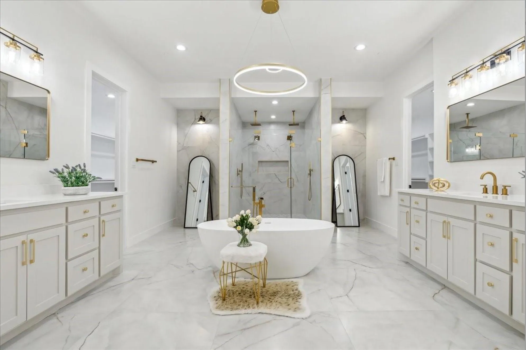 Freestanding soaking tub, dual vanities with white cabinetry, light-toned stone-finish tile flooring, dual shower enclosures with glass doors, and gold-finish fixtures