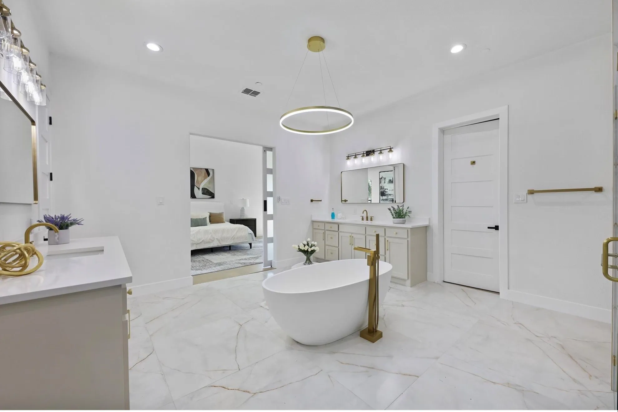 Freestanding soaking tub with a floor-mounted gold-tone faucet, dual vanities with white countertops, marble-look tile flooring, and a contemporary ring chandelier