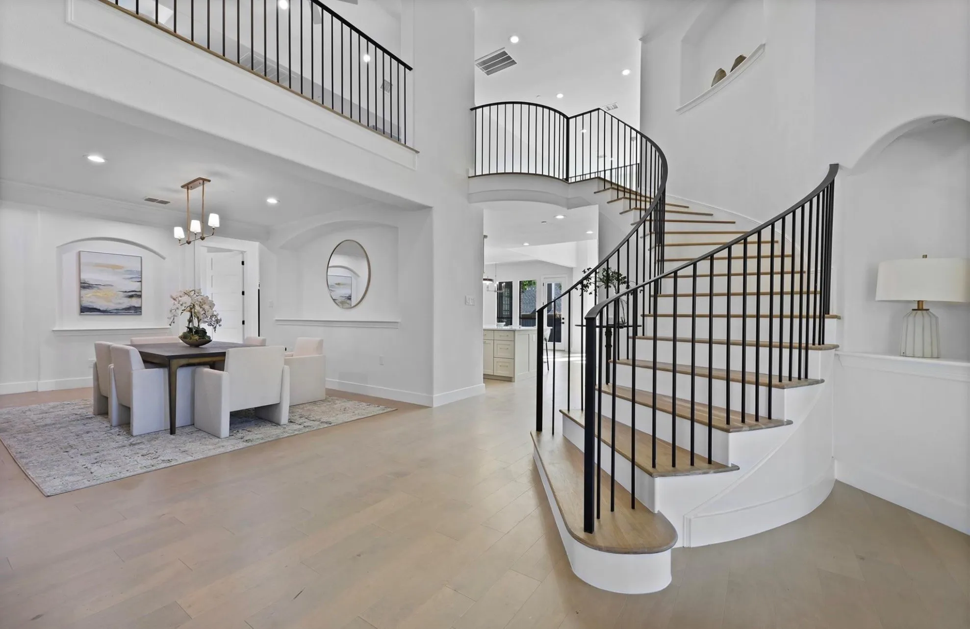 Curved staircase with black wrought iron balusters, light wood-finish flooring, recessed lighting, and an upper-level balcony with matching balusters