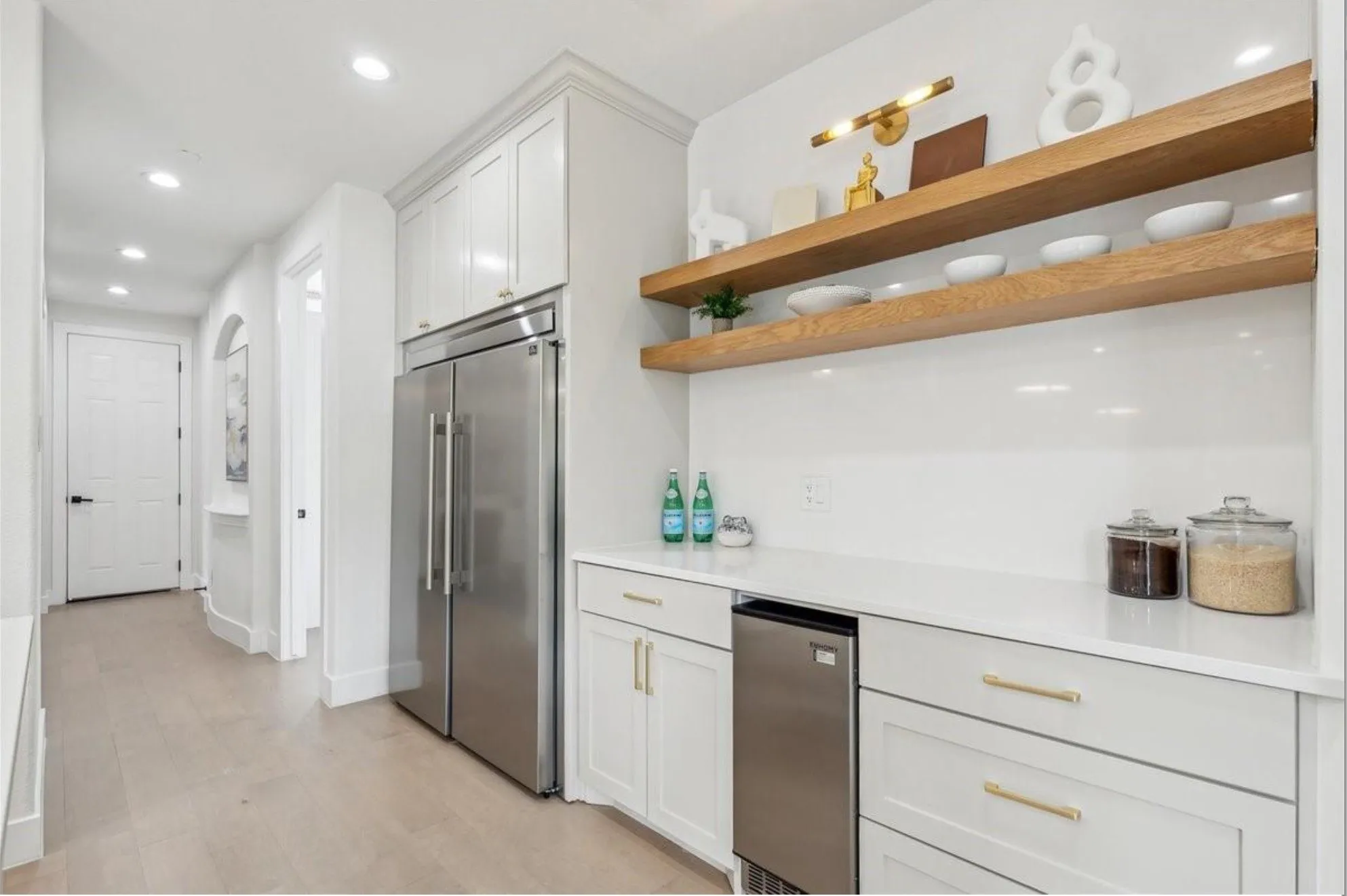 White cabinetry with gold-tone hardware, stainless steel refrigerator, under-counter beverage cooler, white countertop, and wood-finish open shelving