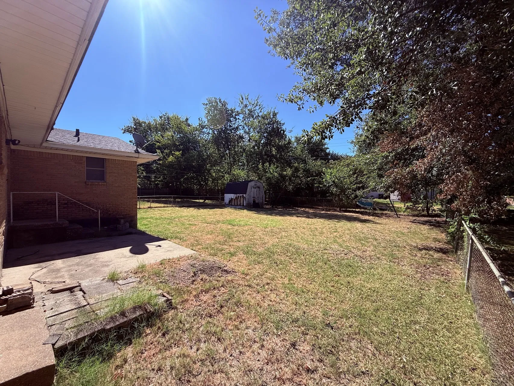 Expansive backyard with a chain-link fence, mature trees, a storage shed, and a concrete patio