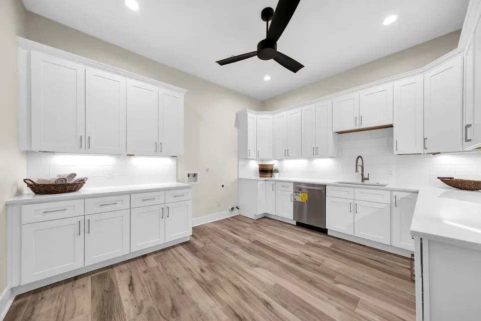 Extensive white cabinetry, integrated stainless steel dishwasher, undermount sink with gooseneck faucet, light wood-finish flooring, and a ceiling fan