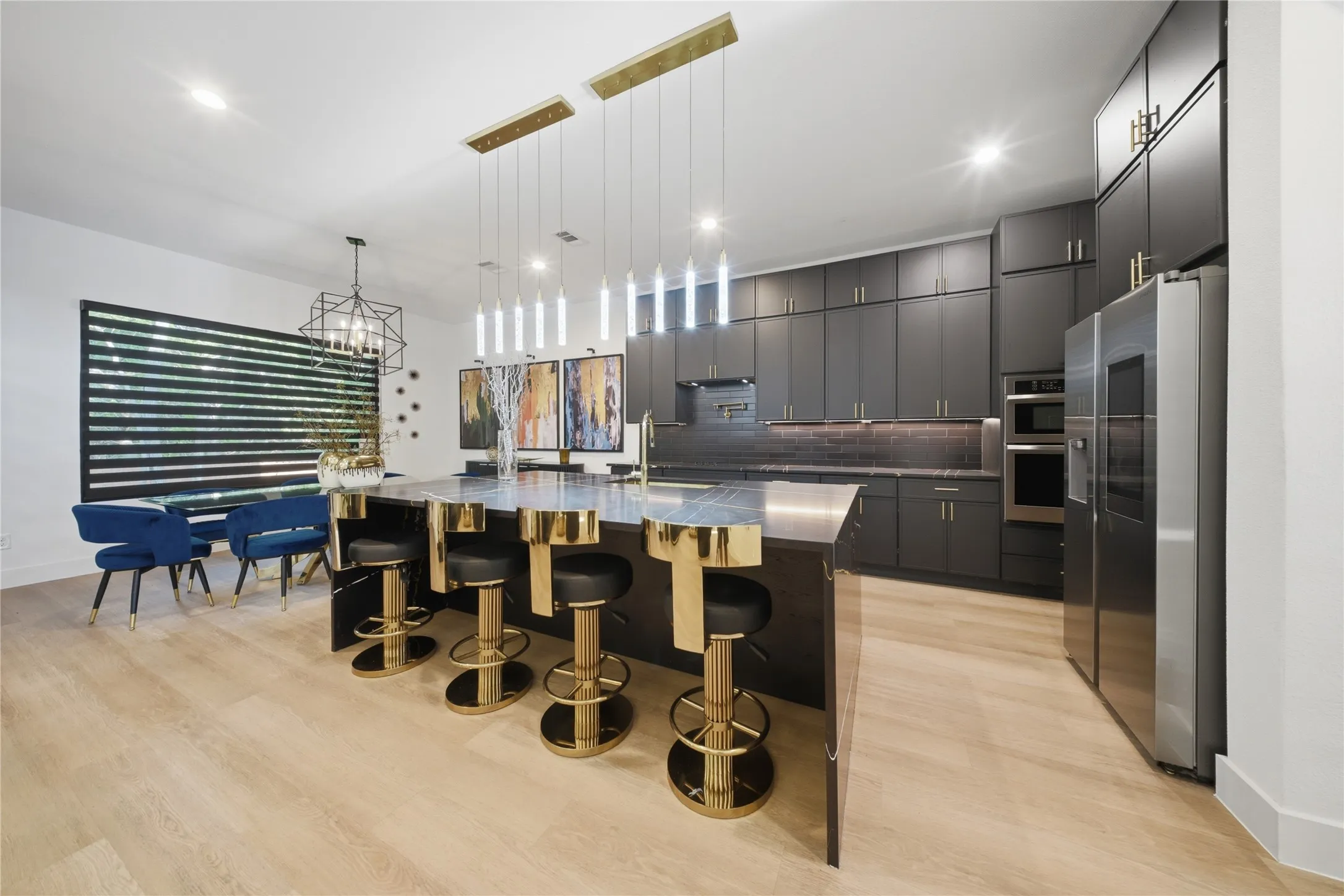 Sleek kitchen featuring dark cabinetry, stainless steel appliances, a large island with pendant lighting, light wood-finish flooring, and a dining area with a geometric chandelier