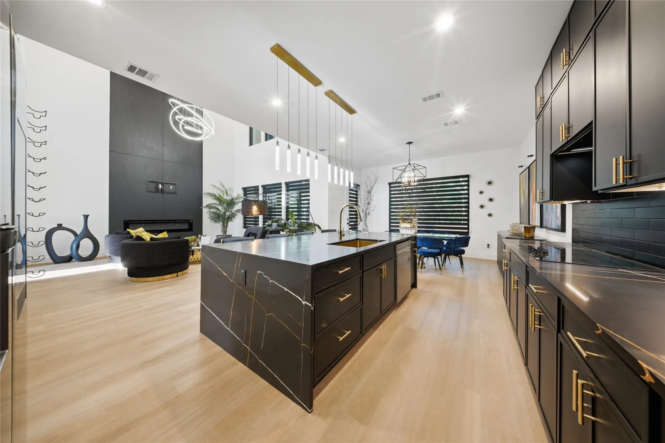 Light wood-finish flooring, dark kitchen cabinetry with gold-tone hardware, a large kitchen island with a stone countertop, a linear pendant light fixture, and a built-in fireplace