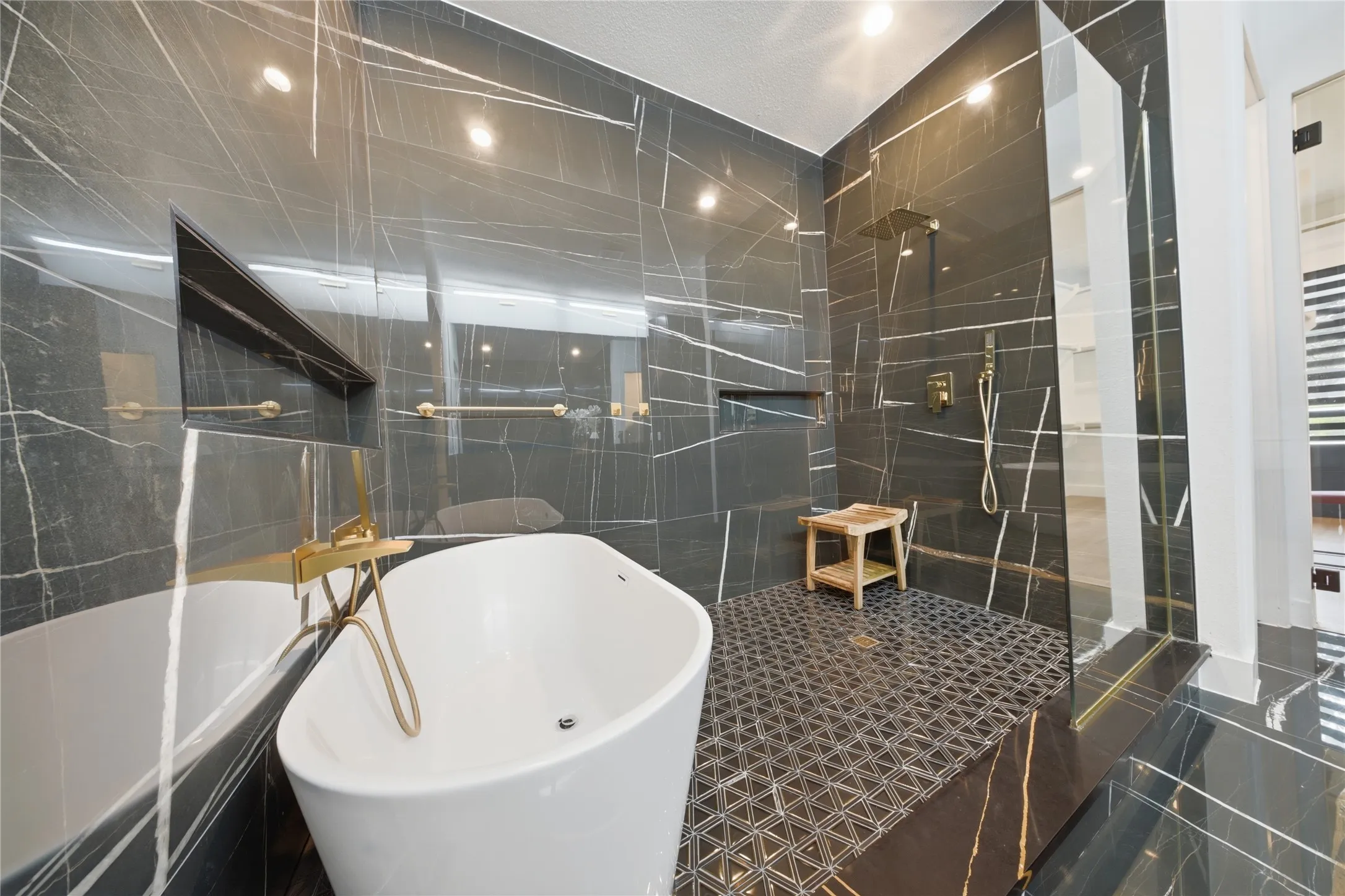 Freestanding soaking tub with a gold-tone floor-mounted faucet, walk-in shower with a glass enclosure, black veined wall tiles, recessed shelving, and a patterned shower floor