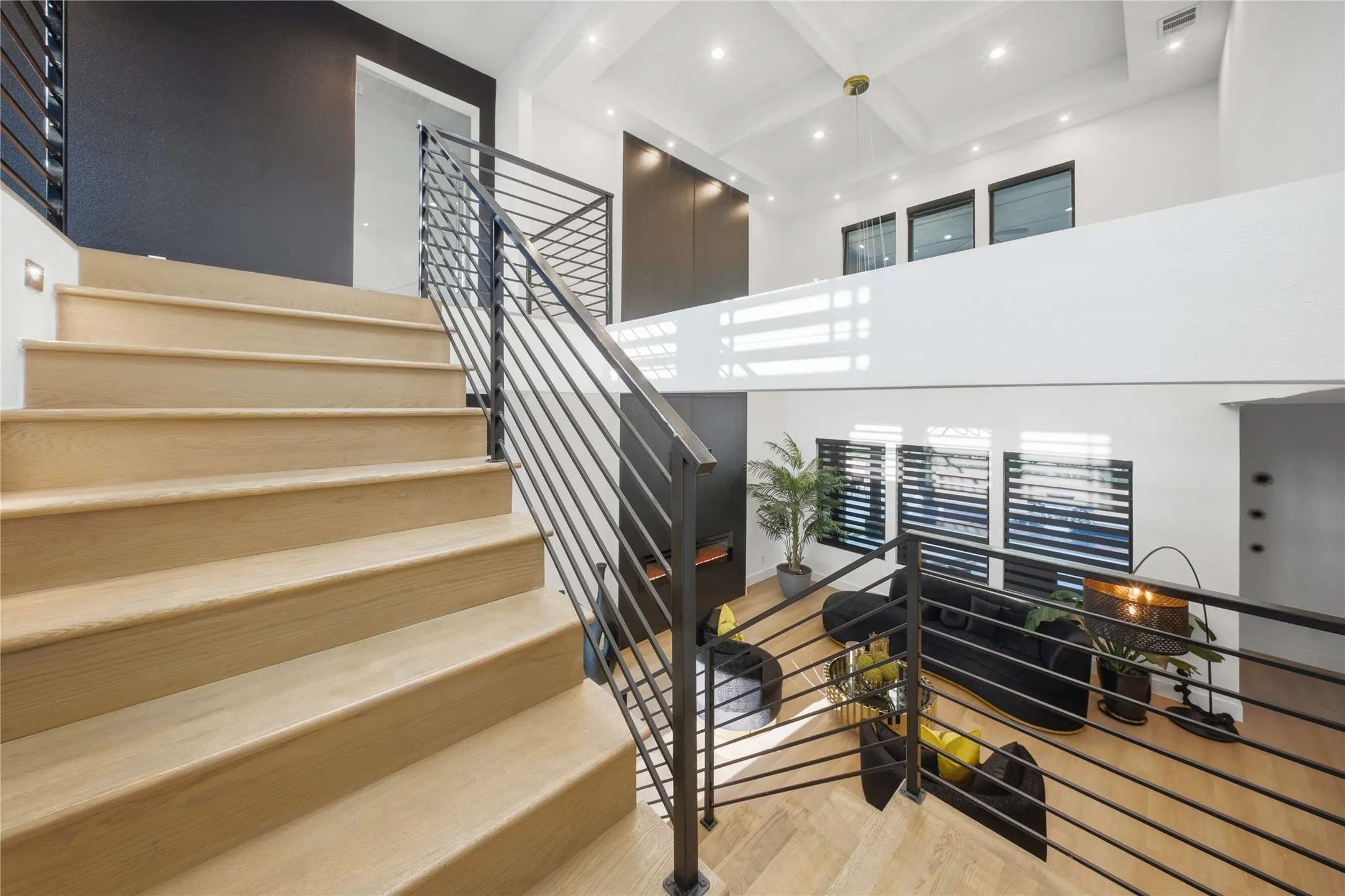 Open-concept living space featuring wood-finish flooring, a modern staircase with metal railings, high ceilings with recessed lighting, and multiple windows with horizontal blinds