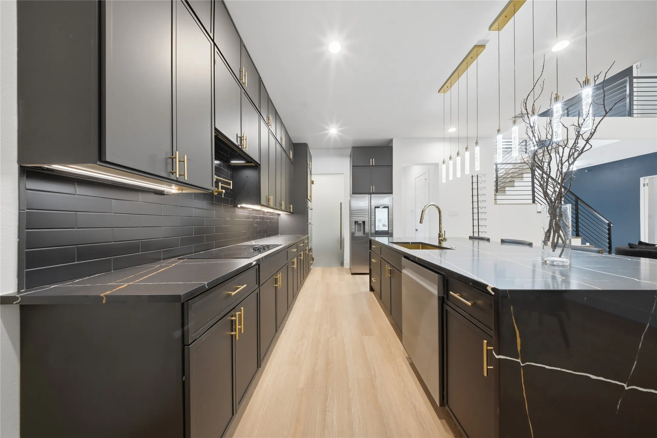 Modern kitchen featuring dark cabinetry with gold-tone hardware, black subway tile backsplash, light wood-finish flooring, and a large island with a gold-veined countertop and integrated sink