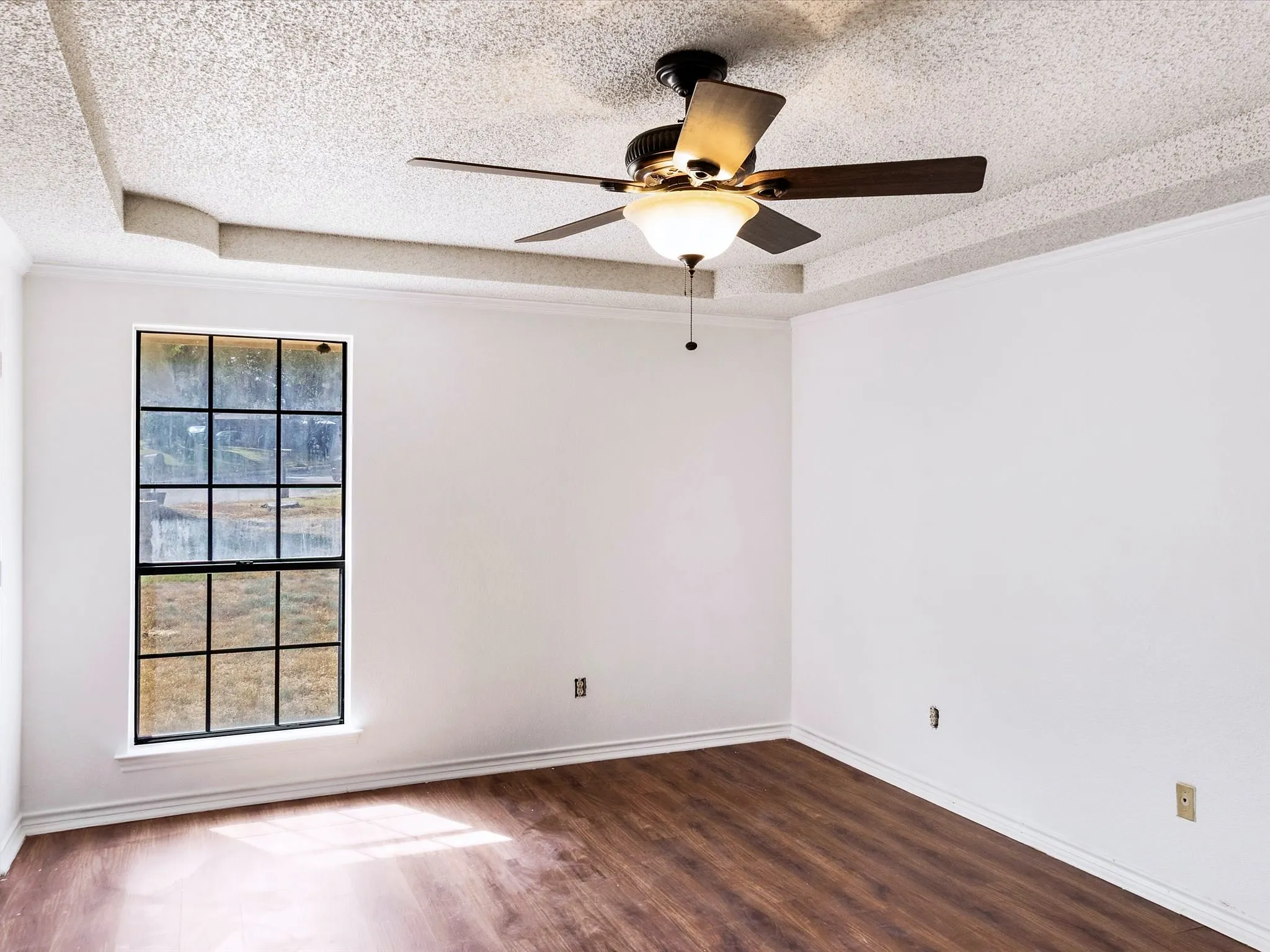 Tray ceiling with textured finish, wood-finish flooring, ceiling fan with integrated lighting, and a multi-pane window