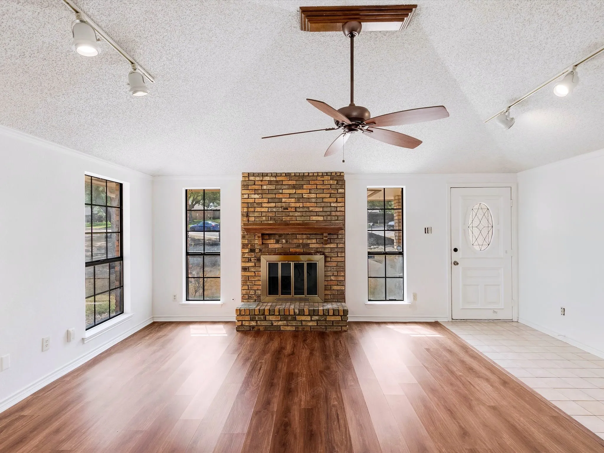 Wood-finish flooring throughout, brick fireplace with wood mantel, ceiling fan, track lighting, and a front entry door with decorative glass inlay