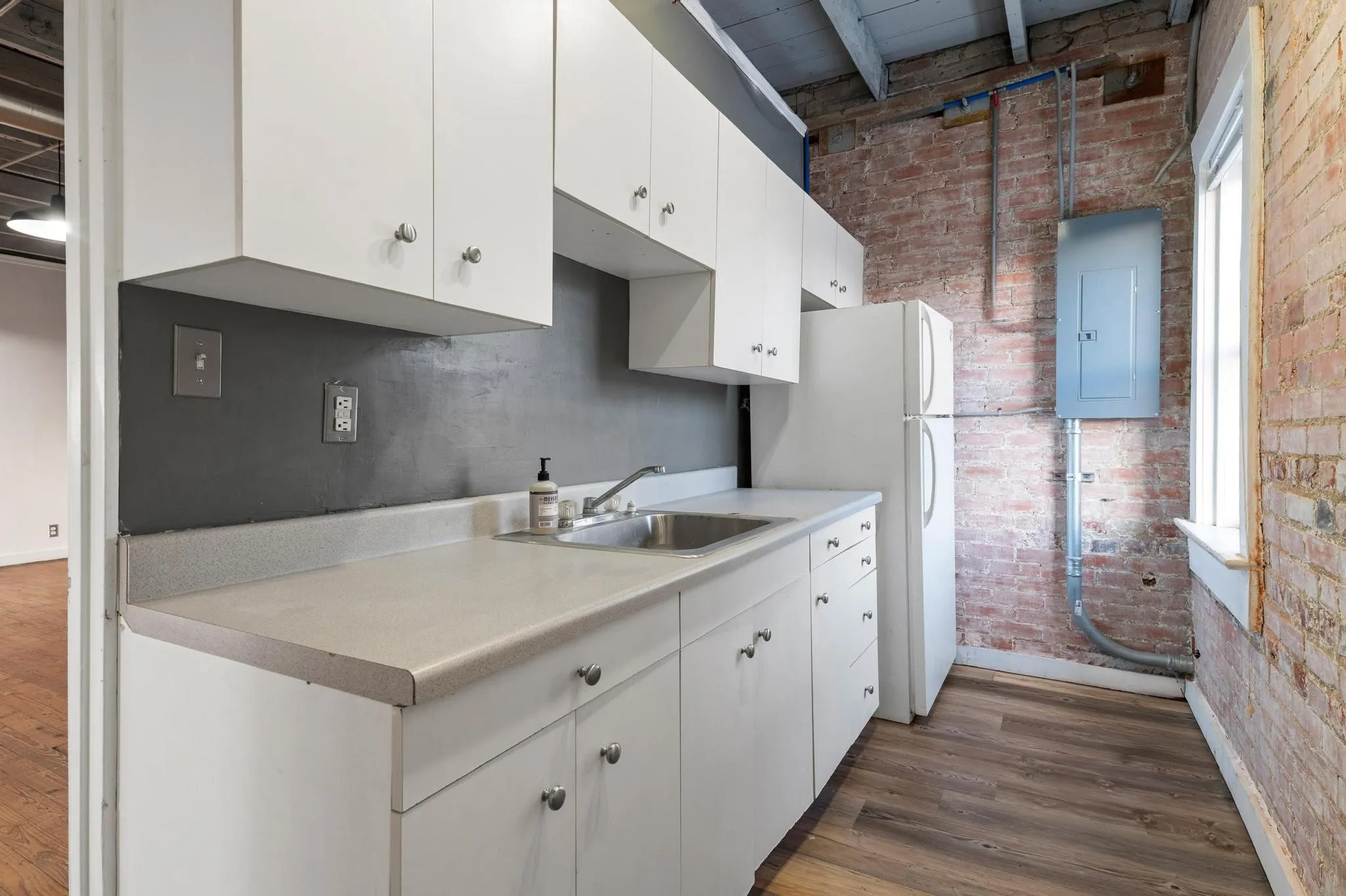 Exposed brick wall, wood-finish flooring, white cabinetry, light-toned countertop, and a window providing natural illumination