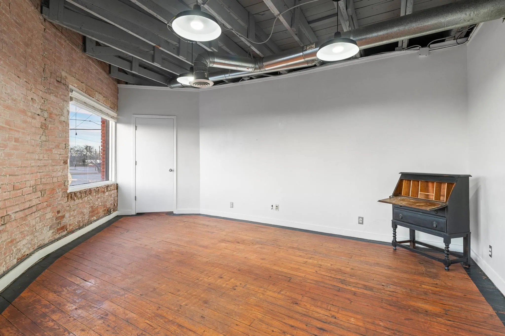 Hardwood flooring, exposed brick wall, overhead industrial lighting, exposed ceiling joists, and a white interior door