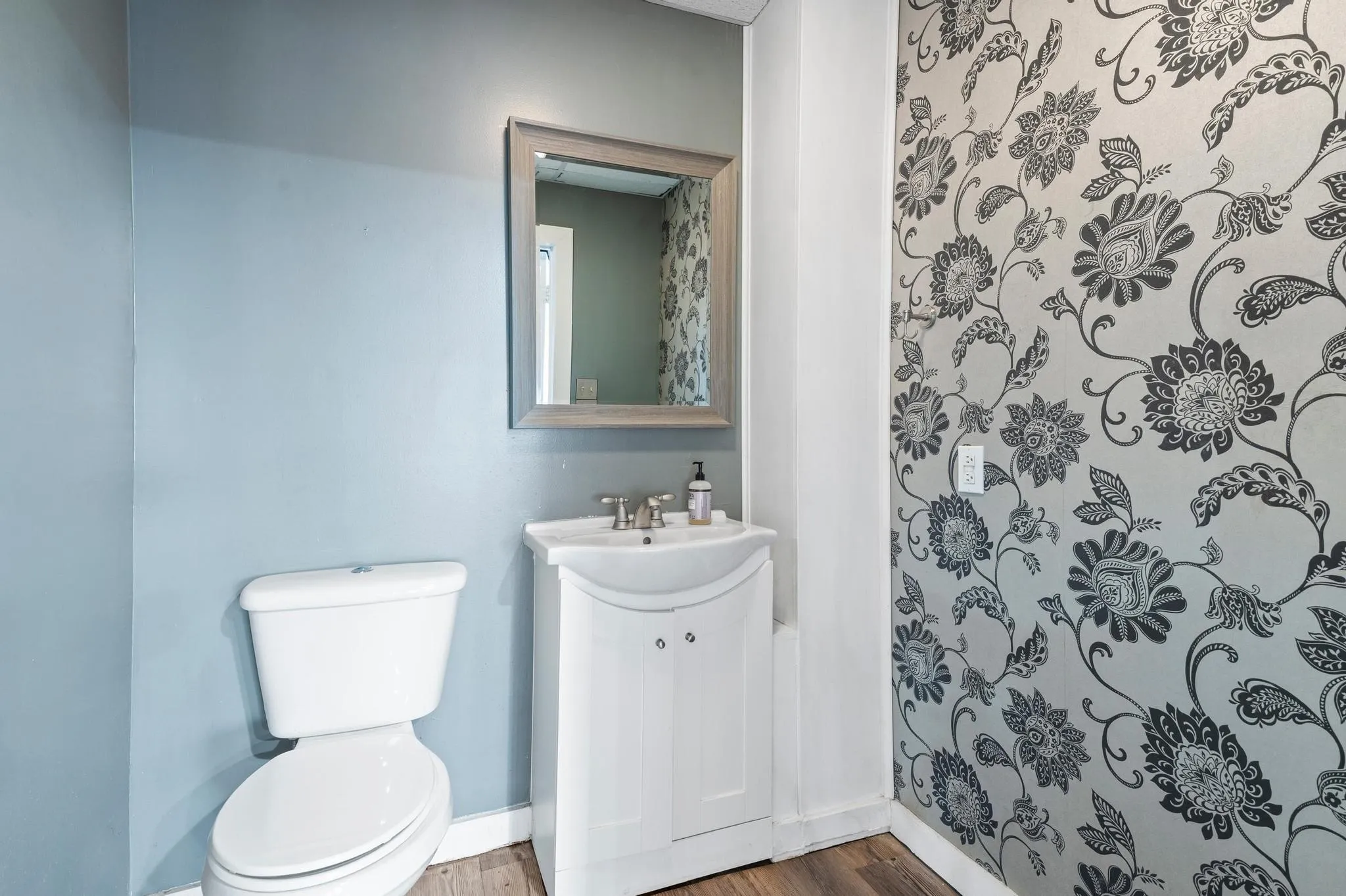 Pedestal sink with a chrome faucet, framed mirror, and decorative floral wallpaper accent wall