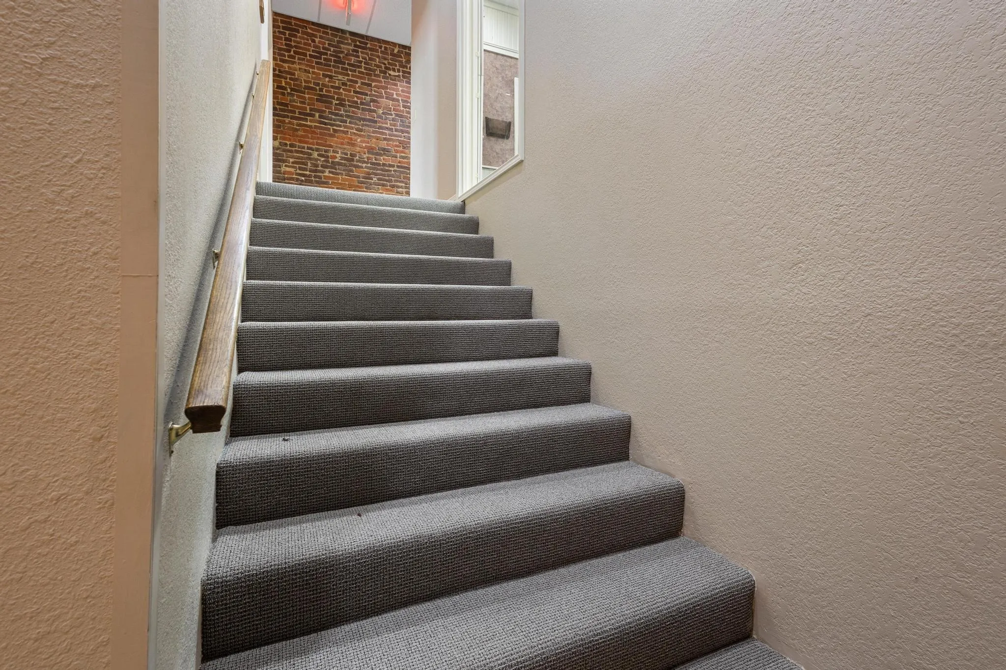 Staircase featuring grey carpeted treads, a wood-finish handrail with brass-tone brackets, and textured wall finishes