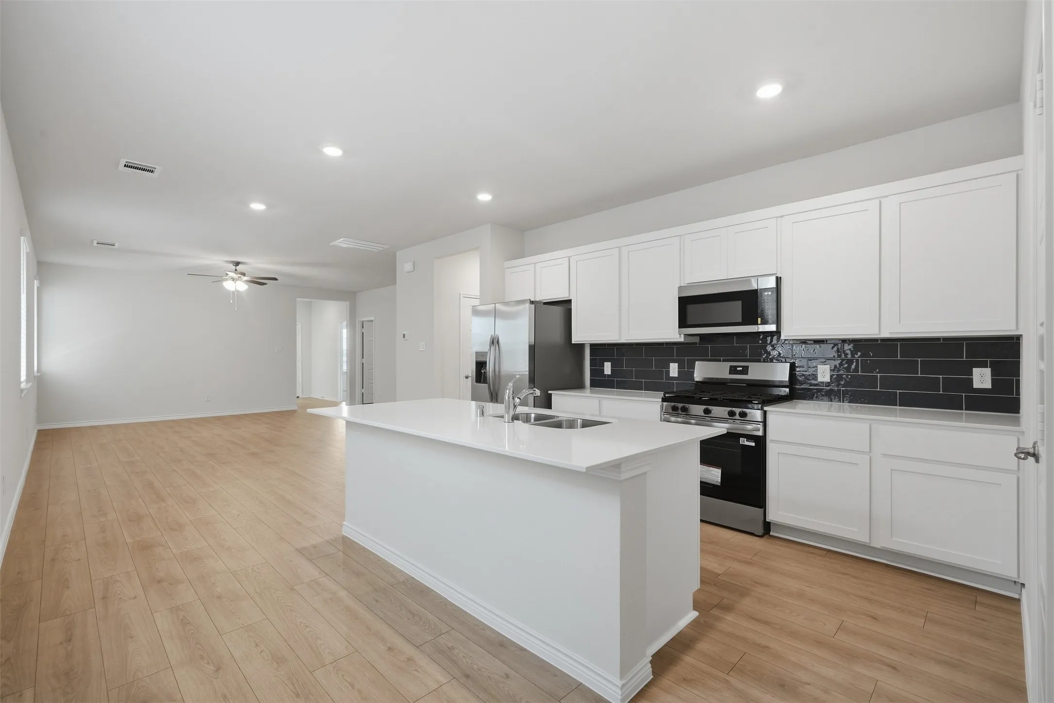 Open-concept kitchen with white shaker cabinetry, stainless steel appliances, dark subway tile backsplash, and a central island with an integrated double sink