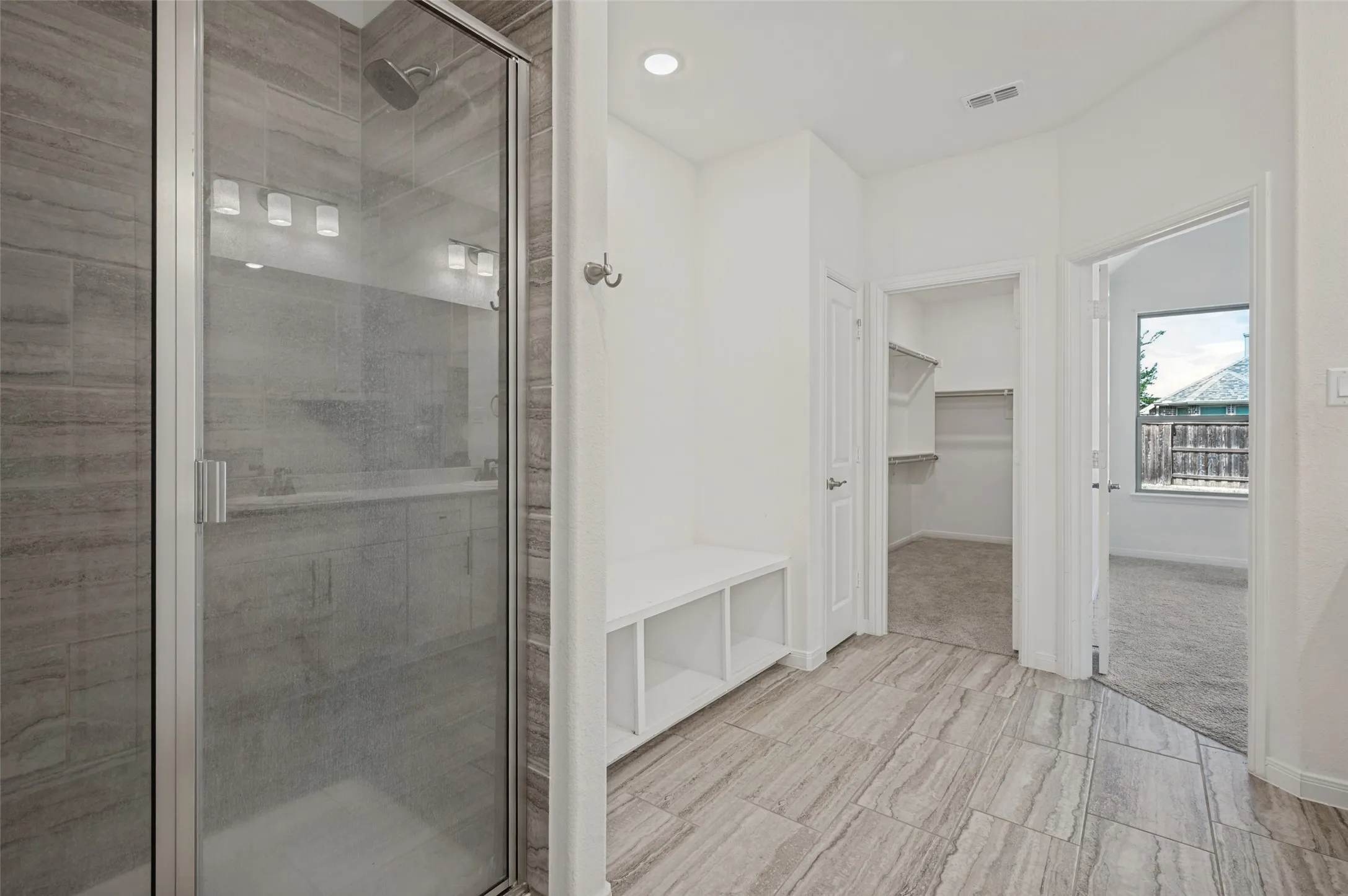 Shower enclosure with glass door and tile surround, white built-in bench with storage, walk-in closet with shelving and hanging rods, light-toned tile flooring, and a window with exterior visibility