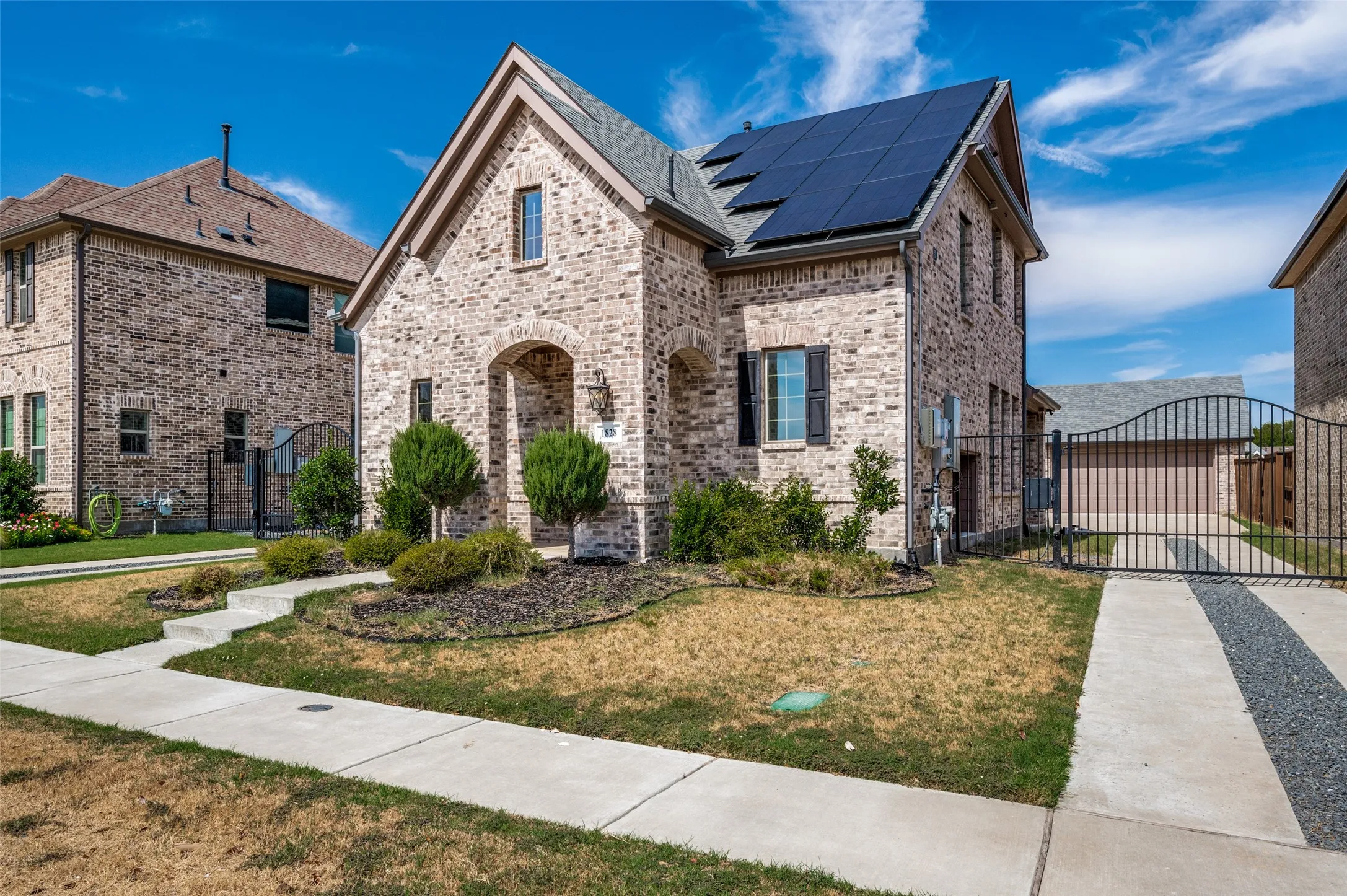 Brick exterior with a gabled roofline, arched entry, solar panel array, and a wrought iron gate leading to a detached garage