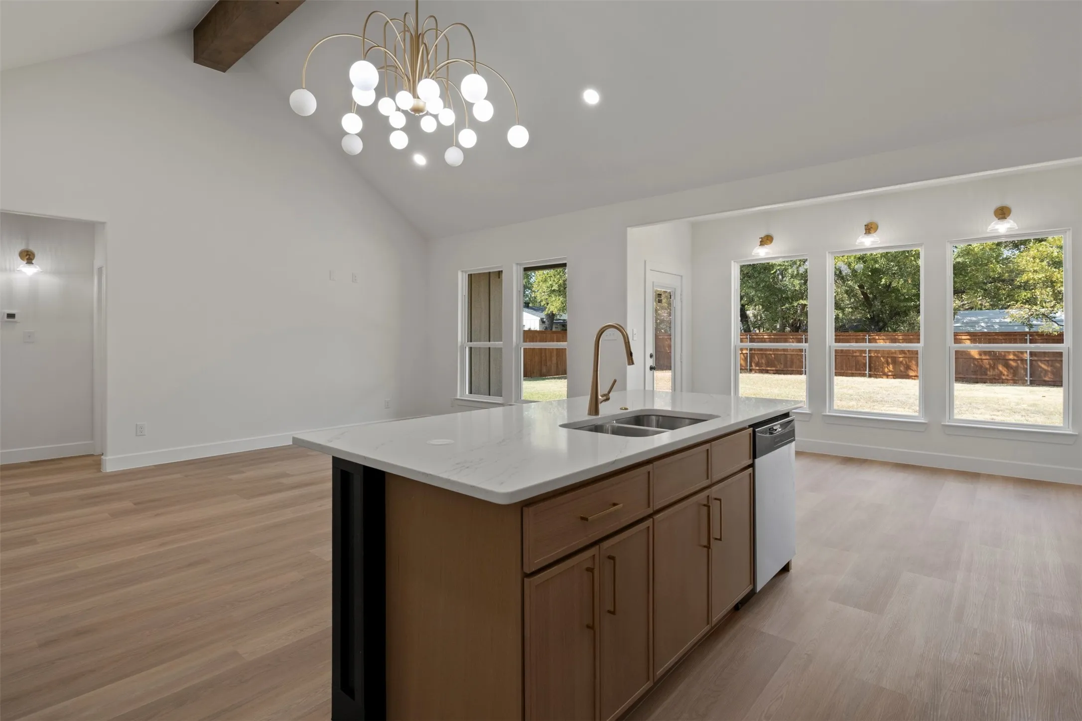 Vaulted ceiling with an exposed wood beam, contemporary chandelier, wood-finish flooring, kitchen island with a double basin sink and integrated dishwasher, and multiple large windows
