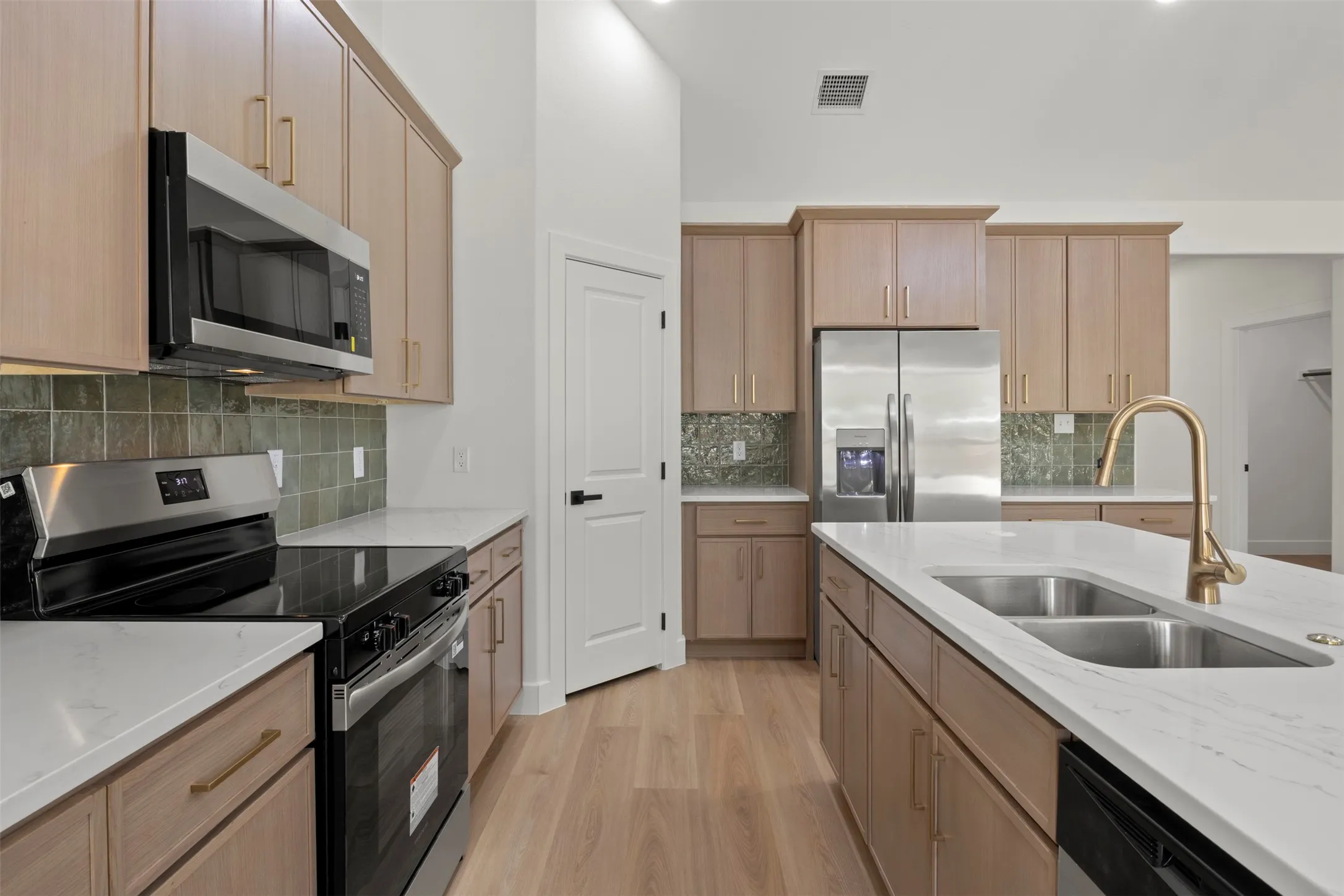 Kitchen featuring wood-finish cabinetry, stainless steel appliances, white countertops, a dual basin sink with a gold-finish faucet, and a green tile backsplash