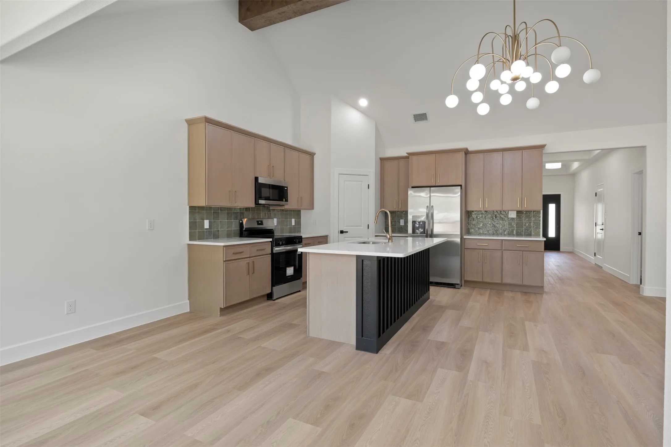 Vaulted ceiling with exposed wood beam, kitchen island with fluted paneling, stainless steel appliances, light wood-finish flooring, and modern chandelier