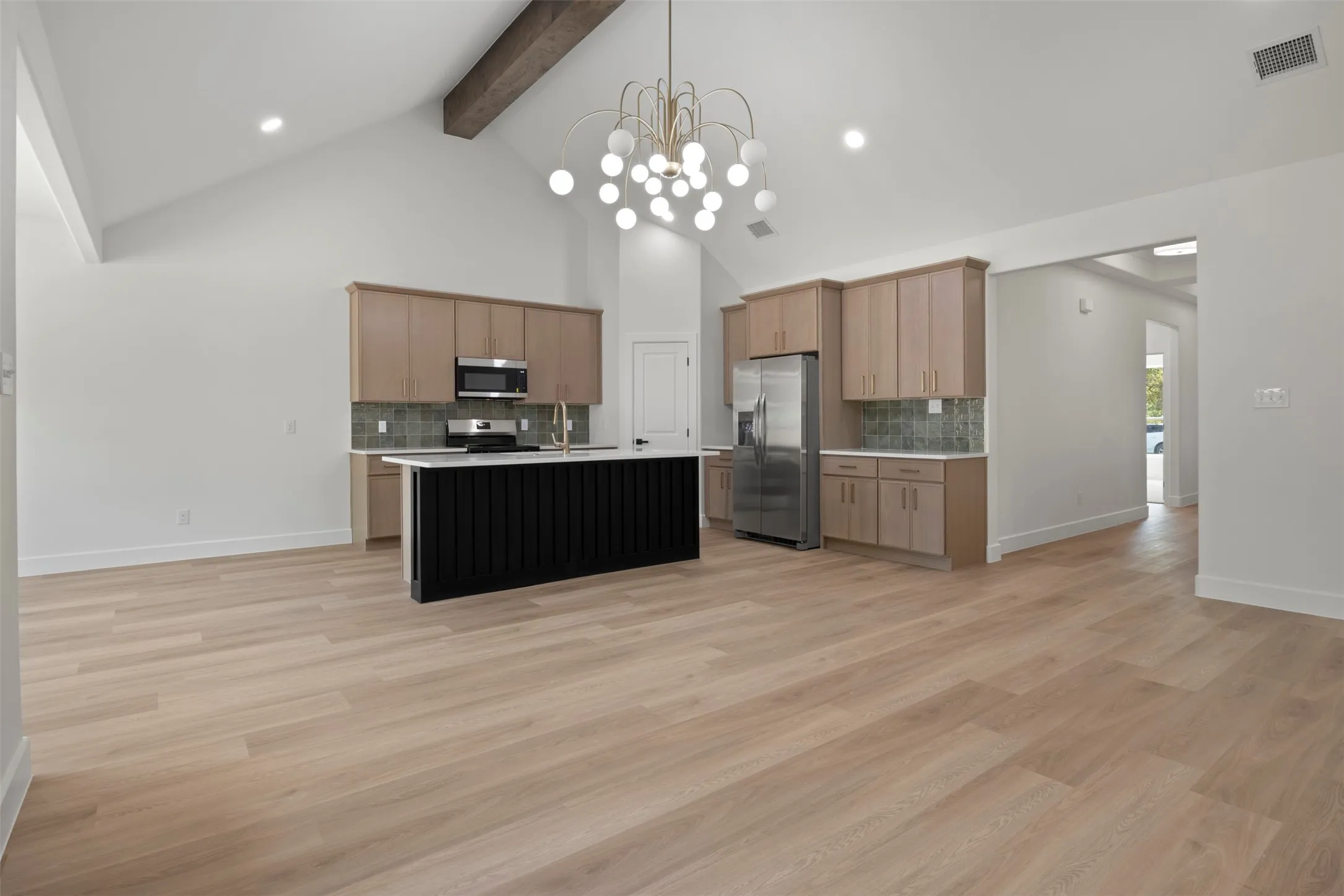 Vaulted ceiling with exposed wood beam, wood-finish flooring, kitchen island with dark paneling, light wood cabinetry, and stainless steel appliances