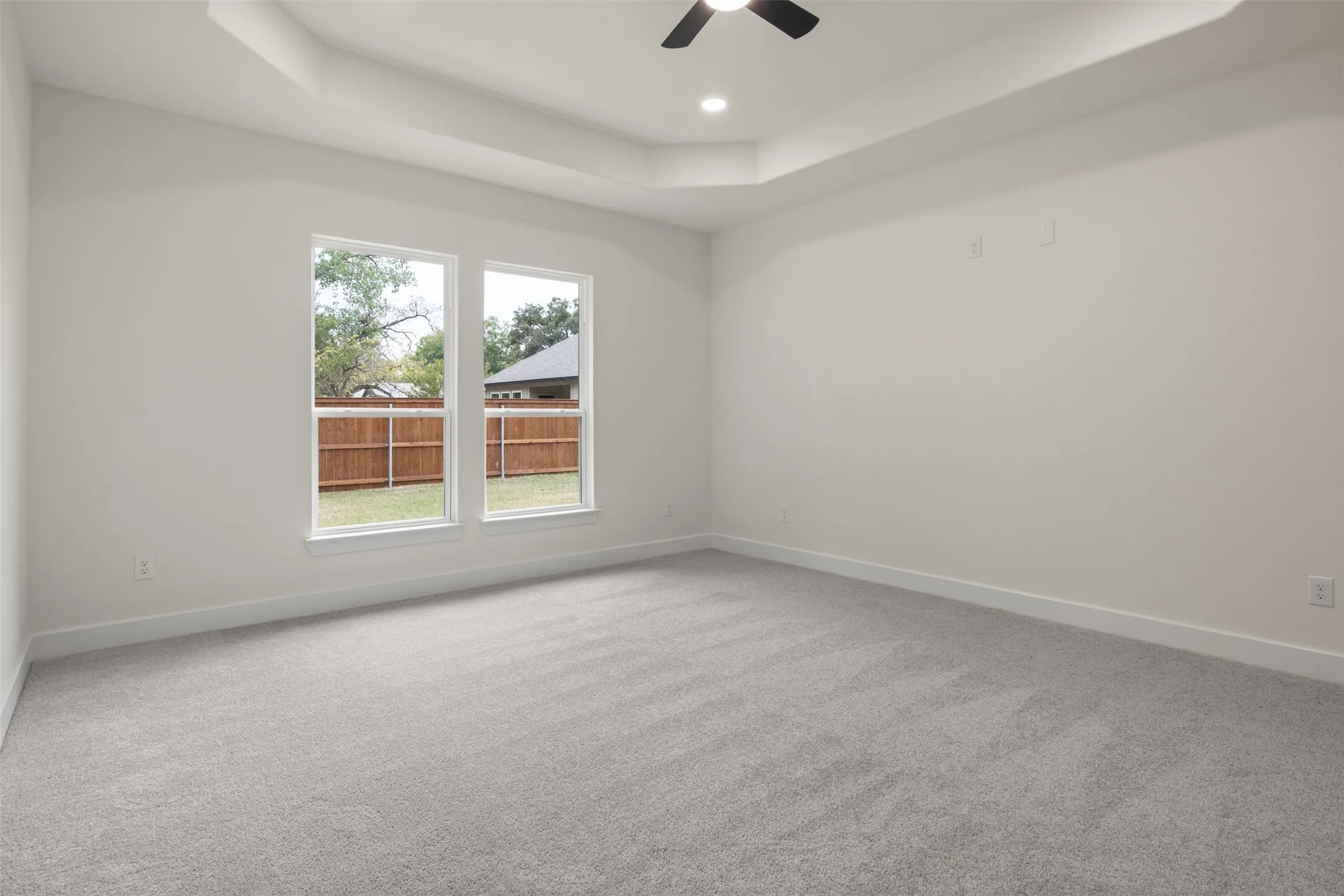 Tray ceiling with recessed lighting and a ceiling fan, two large windows, and neutral carpet