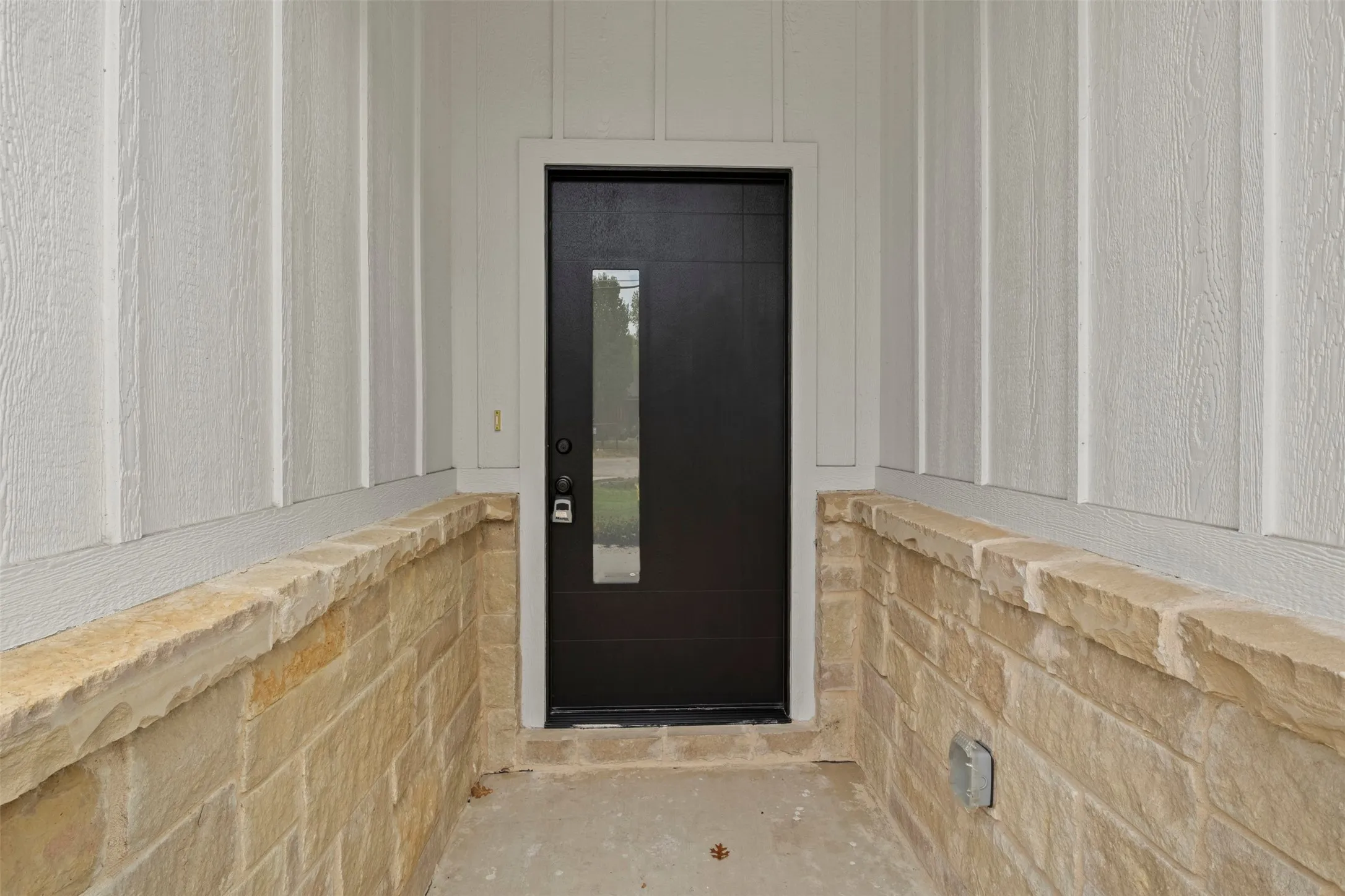 Board and batten siding with a stone veneer foundation and a contemporary dark entry door featuring a vertical privacy glass insert