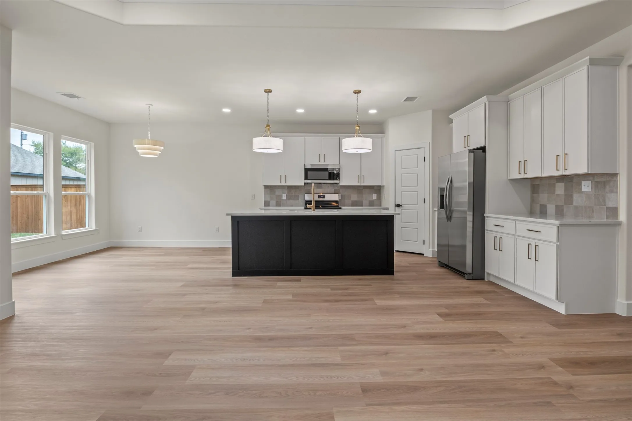 Spacious kitchen and living area featuring wood-finish flooring, white cabinetry with gold-tone hardware, a black kitchen island, stainless steel appliances, and ample natural light from multiple windows