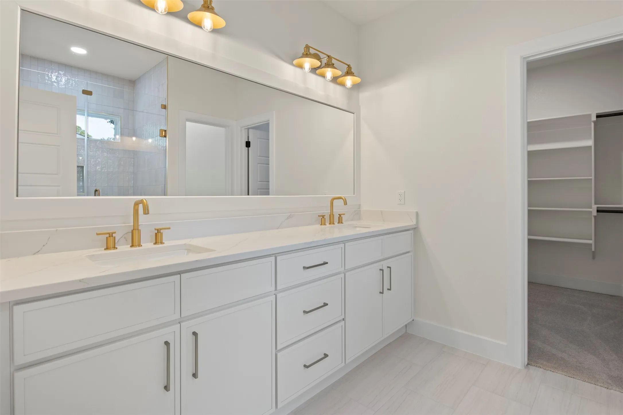 Dual vanity with gold-finish faucets and hardware, white cabinetry, a full-width mirror, and tile flooring