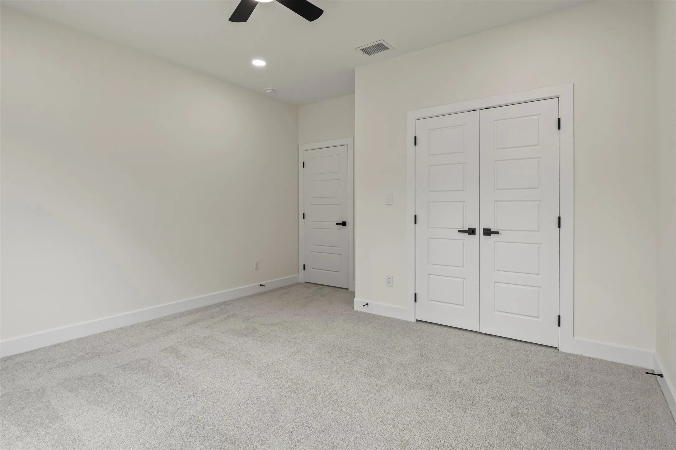 Light gray carpet flooring, white walls, two paneled doors with matte black hardware, and a ceiling fan with integrated lighting