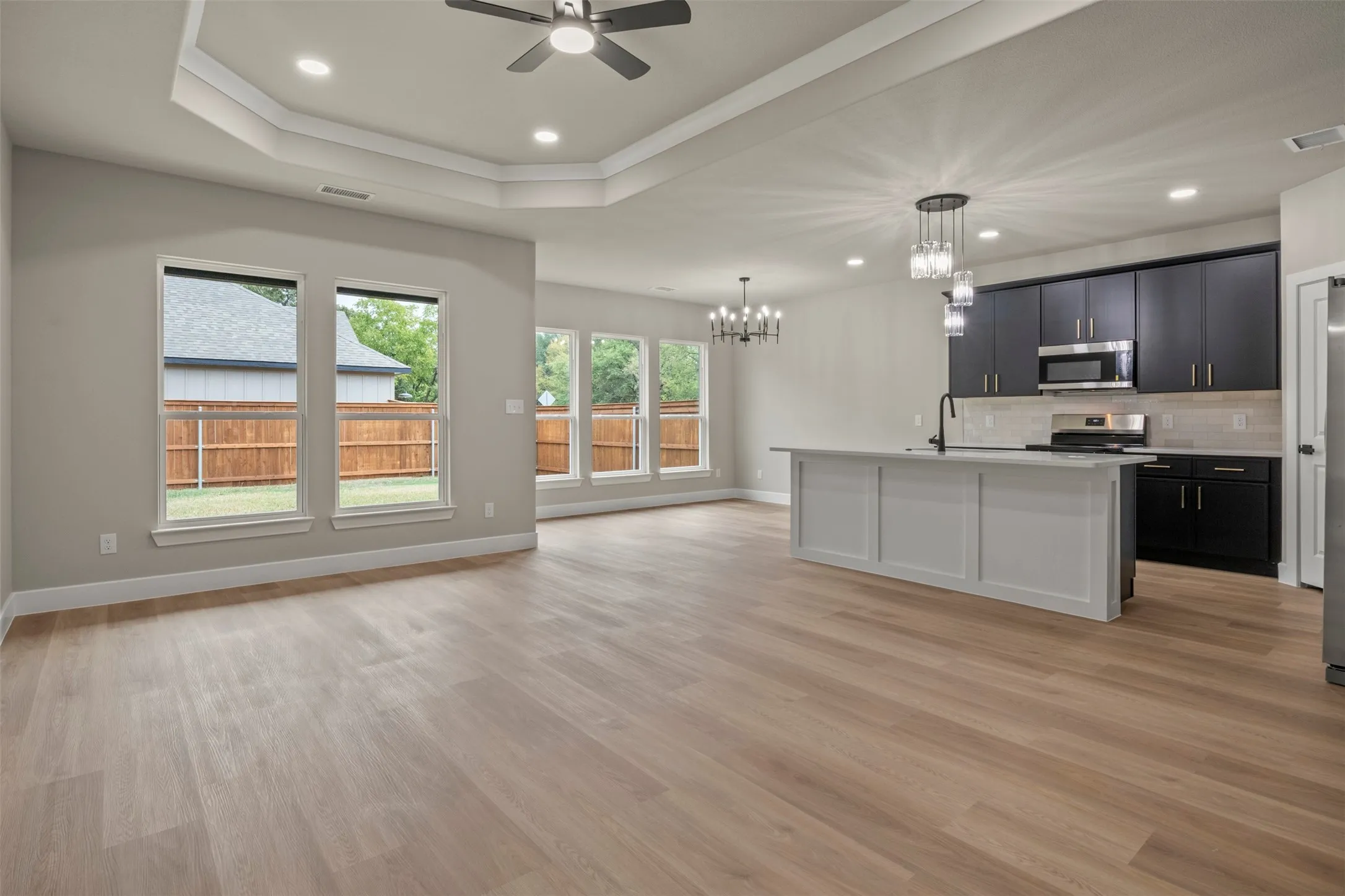 Open-concept living space featuring wood-finish flooring, a kitchen island with pendant lighting, black cabinetry, and a tray ceiling with a ceiling fan
