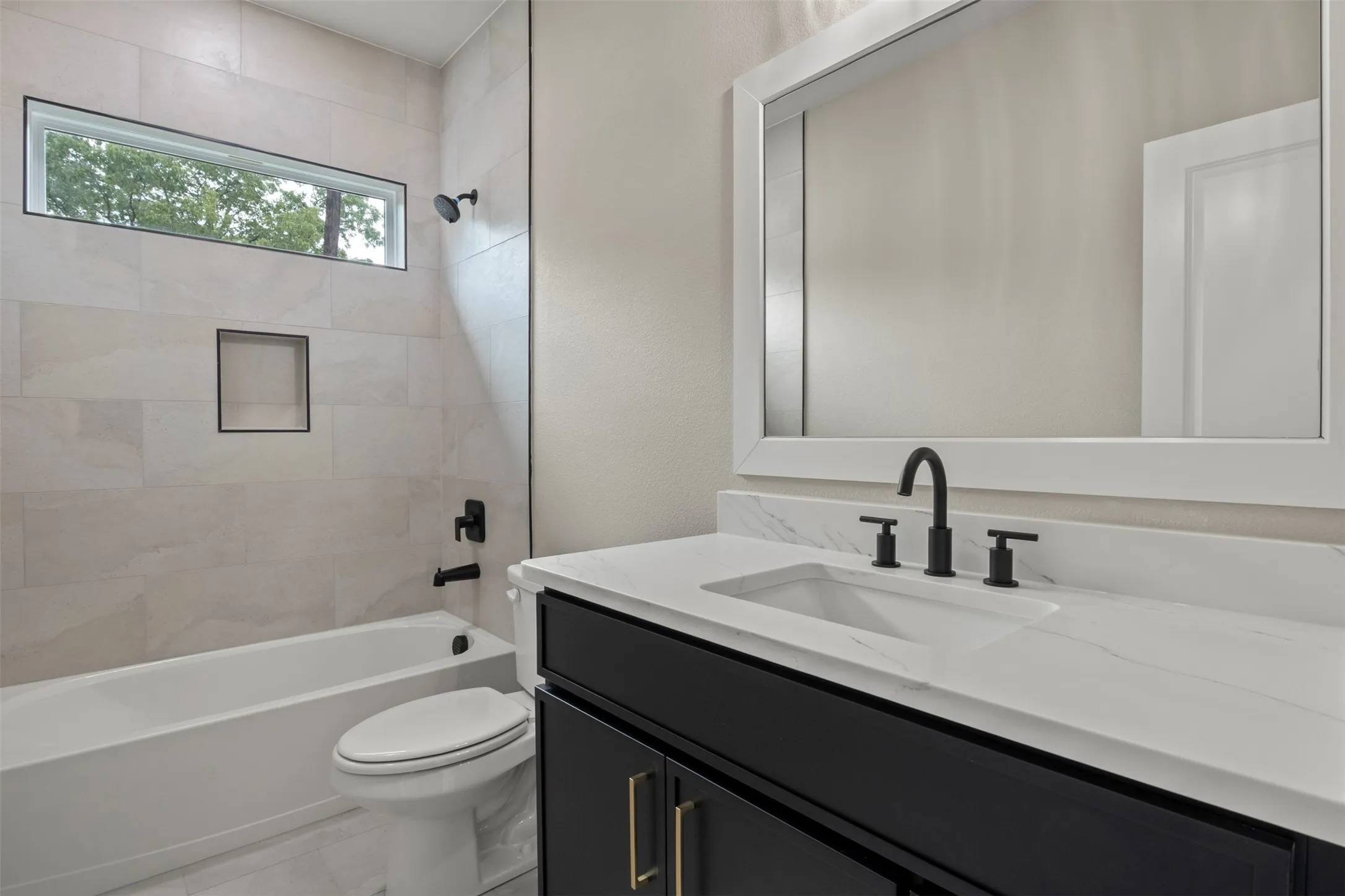 White rectangular sink with matte black faucet, dark wood-finish vanity with gold-tone hardware, white framed mirror, and tiled shower surround with a built-in niche