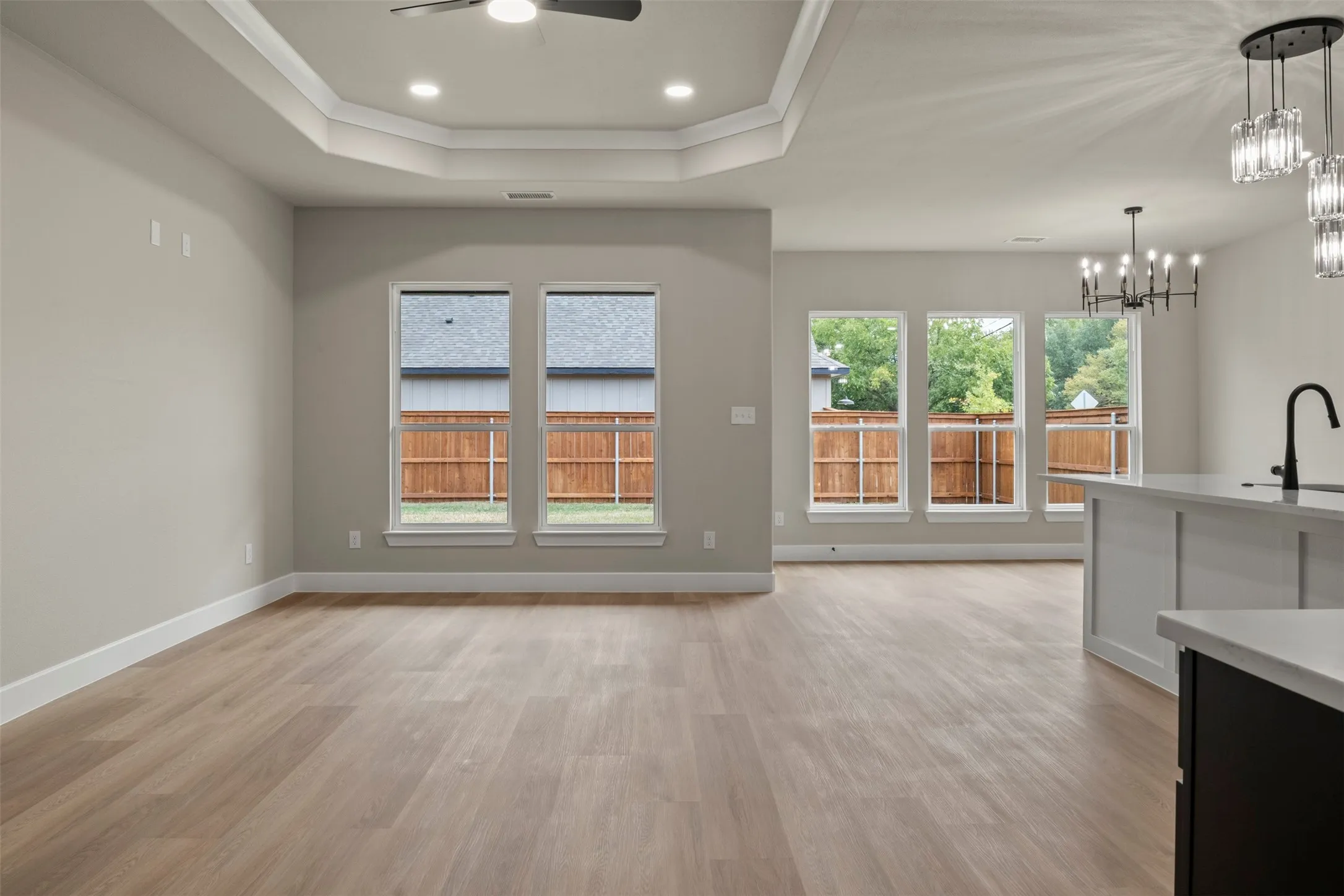 Spacious living area featuring wood-finish flooring, a tray ceiling with recessed lighting, multiple windows, and a built-in kitchen island with a matte black faucet