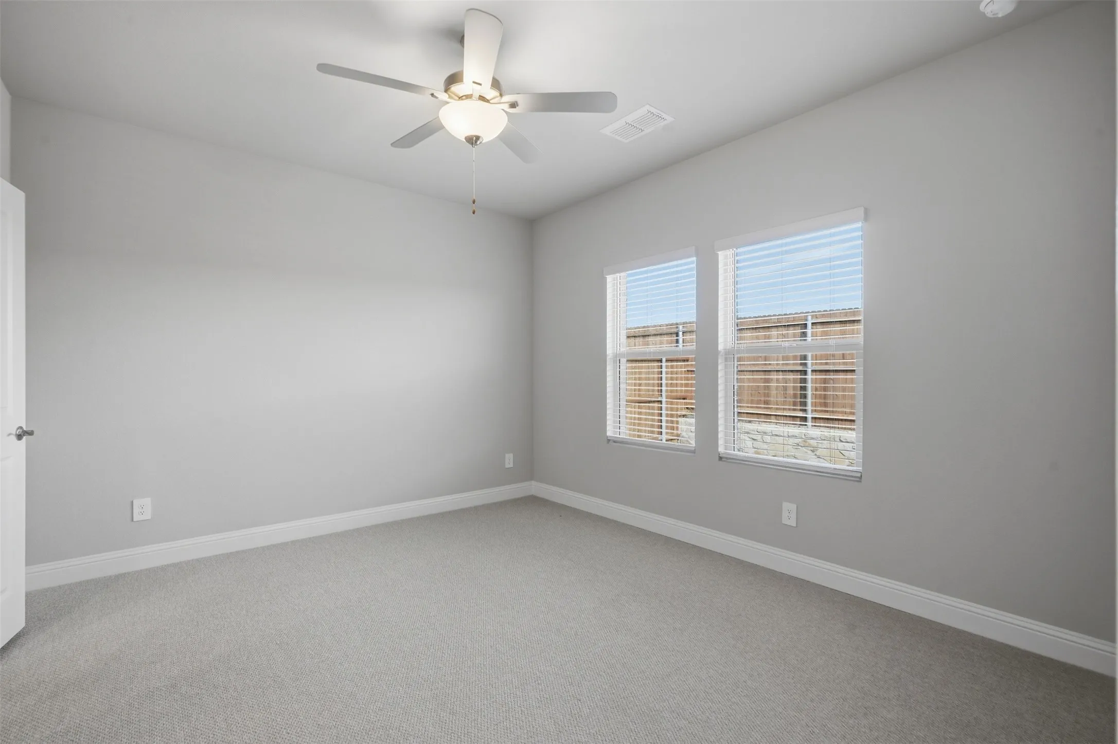 Neutral tone wall paint, light gray carpet, ceiling fan with integrated lighting, two windows with blinds, and white trim