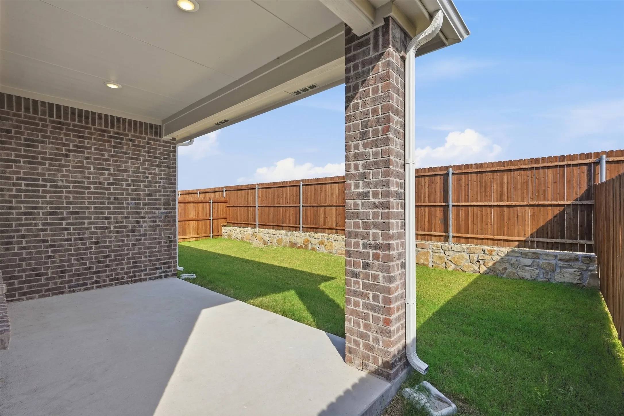 Covered patio with polished concrete flooring and brick-veneer support column, opening to a level lawn with wood privacy fencing and mature landscaping