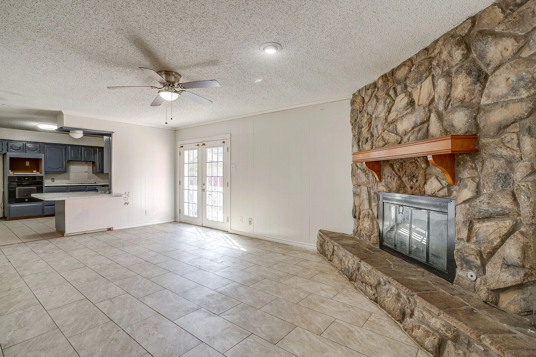 Stone fireplace with wood mantel, tile flooring, built-in kitchen cabinetry, and French patio doors