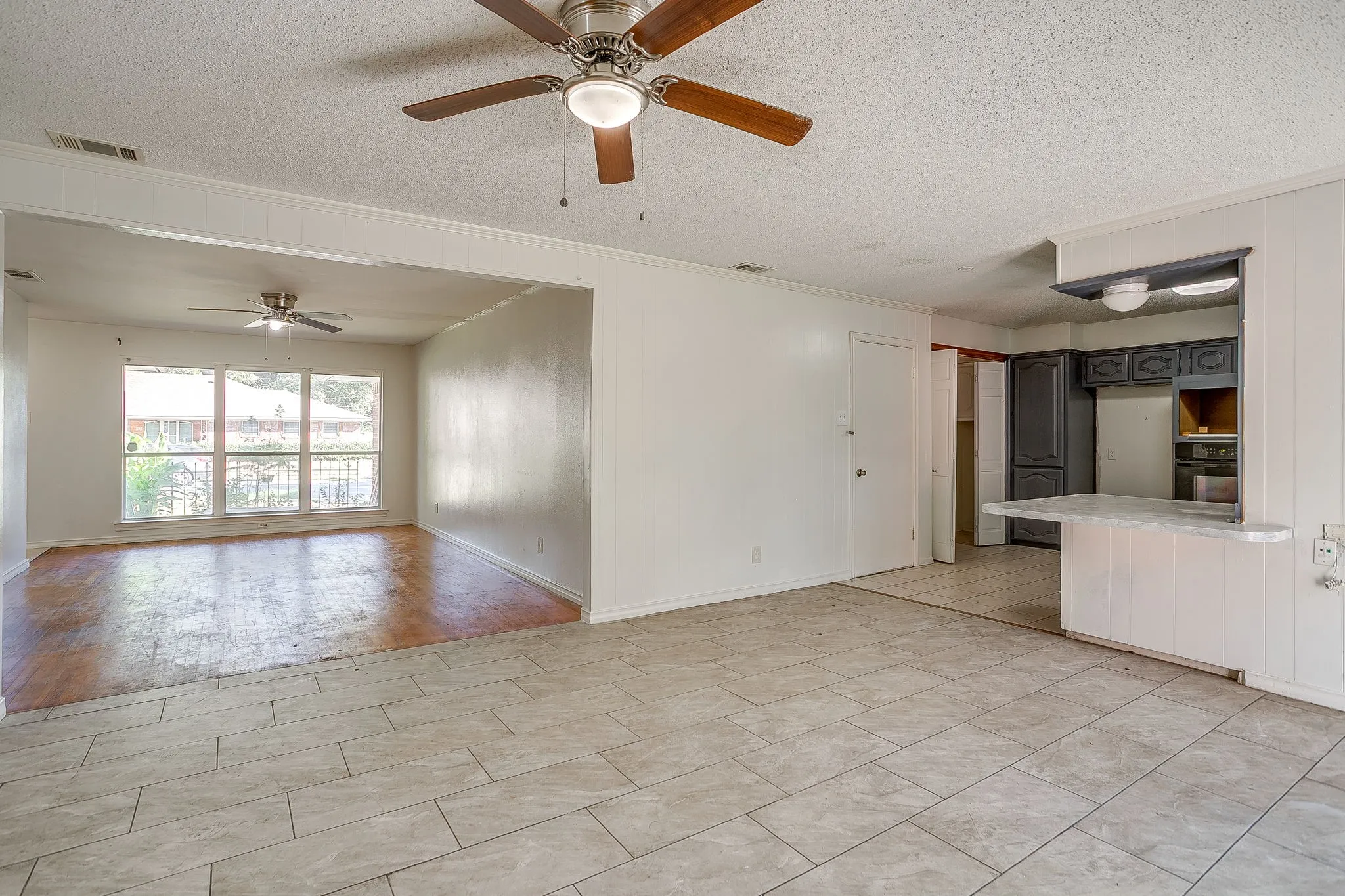 Expansive room featuring light-toned tile flooring, a ceiling fan with integrated lighting, and a pass-through counter with dark cabinetry