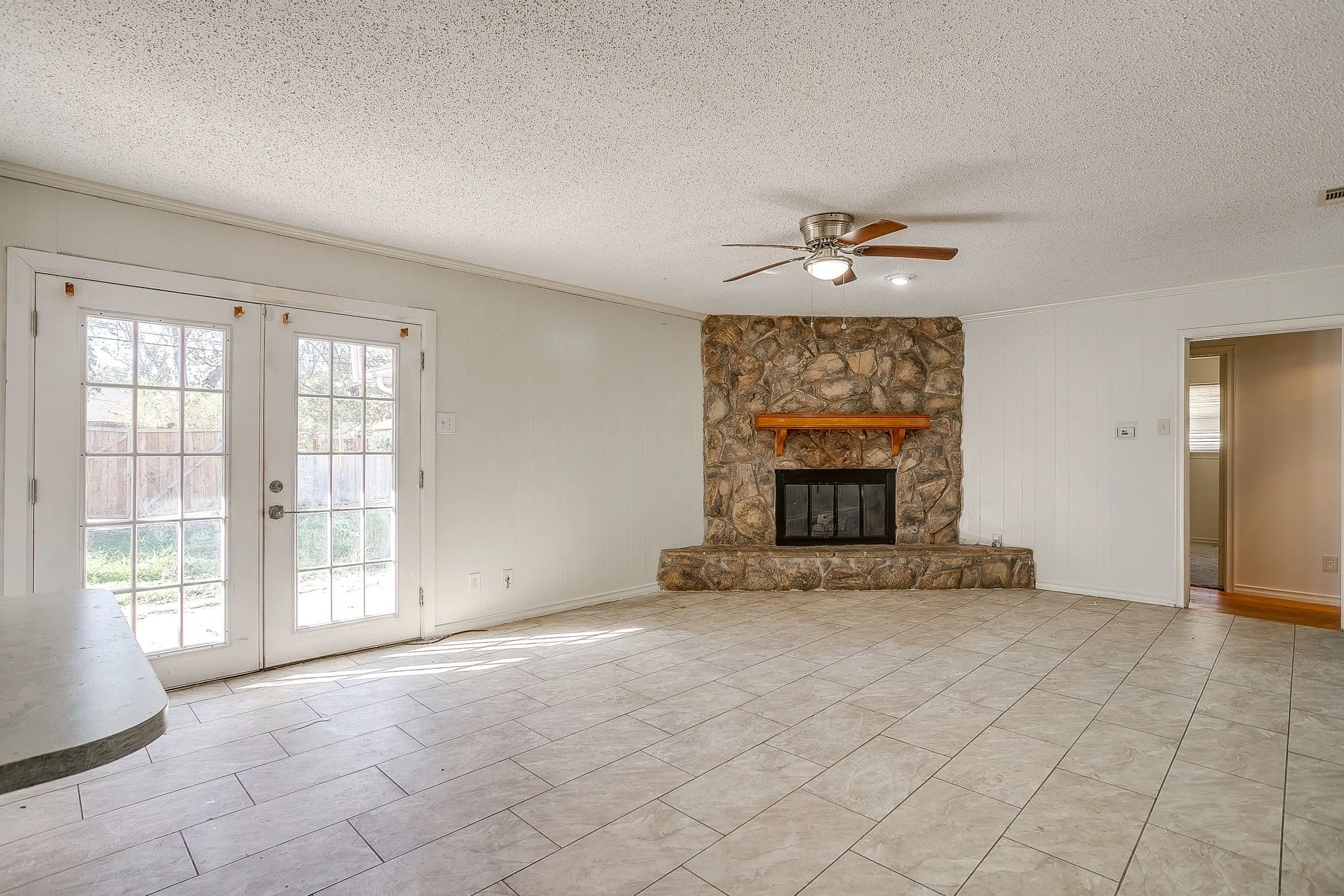 Tile flooring, stone fireplace with wood mantel, ceiling fan with integrated lighting, white French doors, and a partial breakfast bar