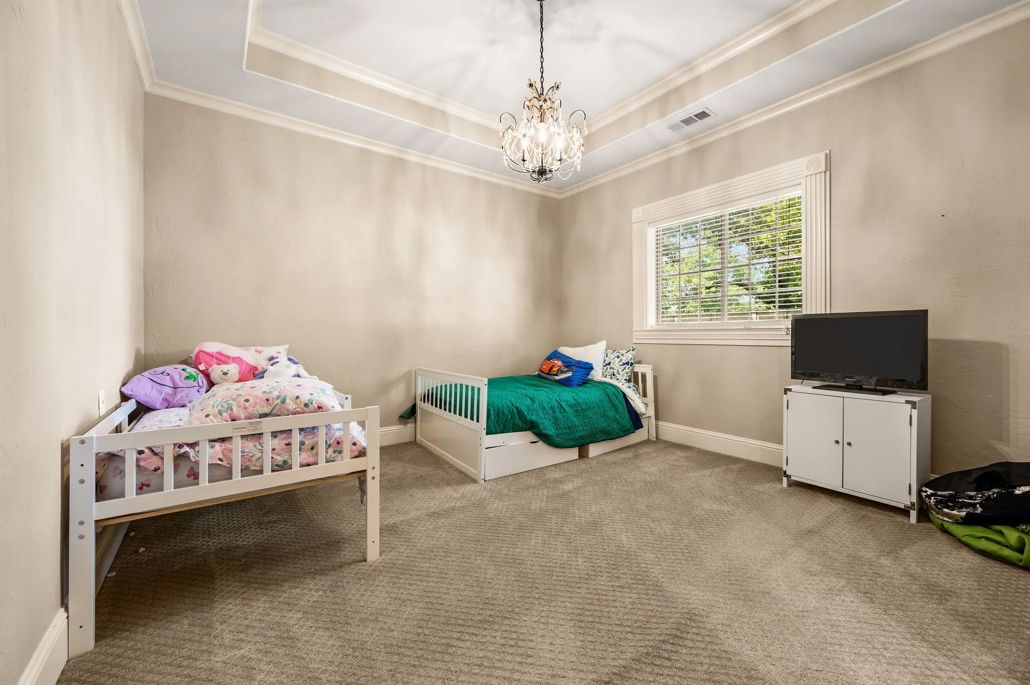 Recessed ceiling with an ornate chandelier, window with white blinds, and light-toned carpeting