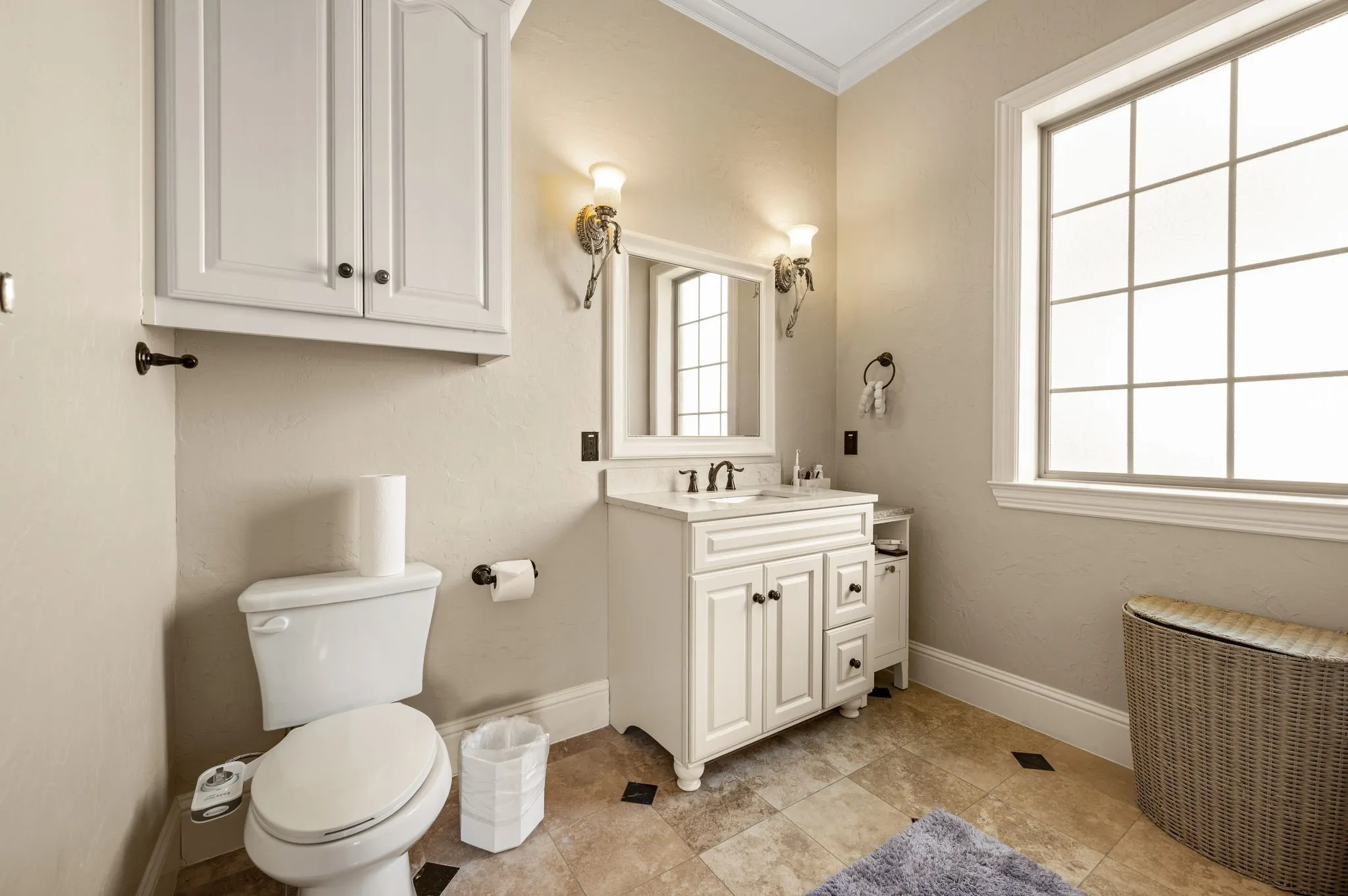 White vanity with undermount sink, wall-mounted sconce lighting, an overhead cabinet, and a large window